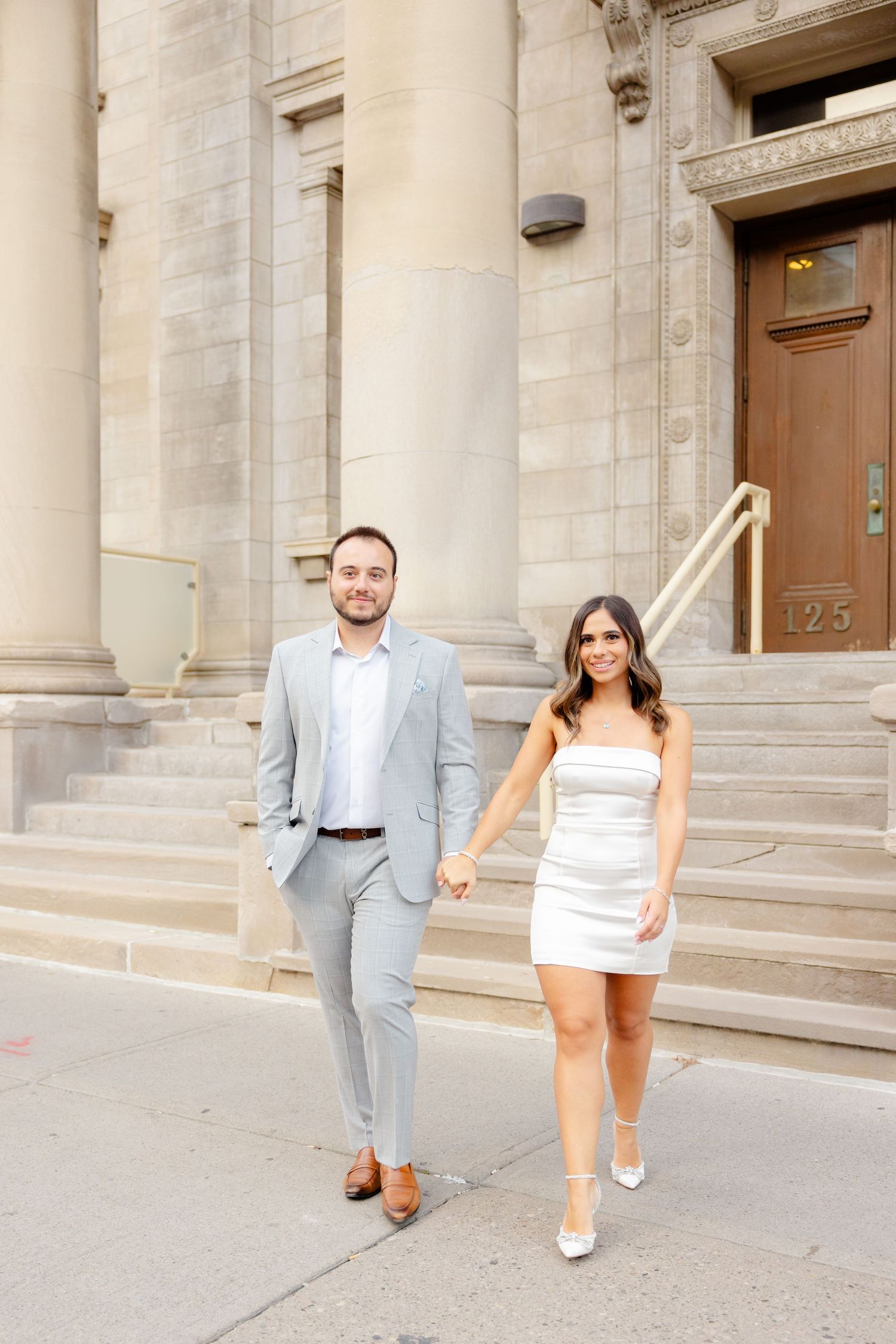 A stylish couple in coordinating formal wear walks hand in hand outside a historic building.
