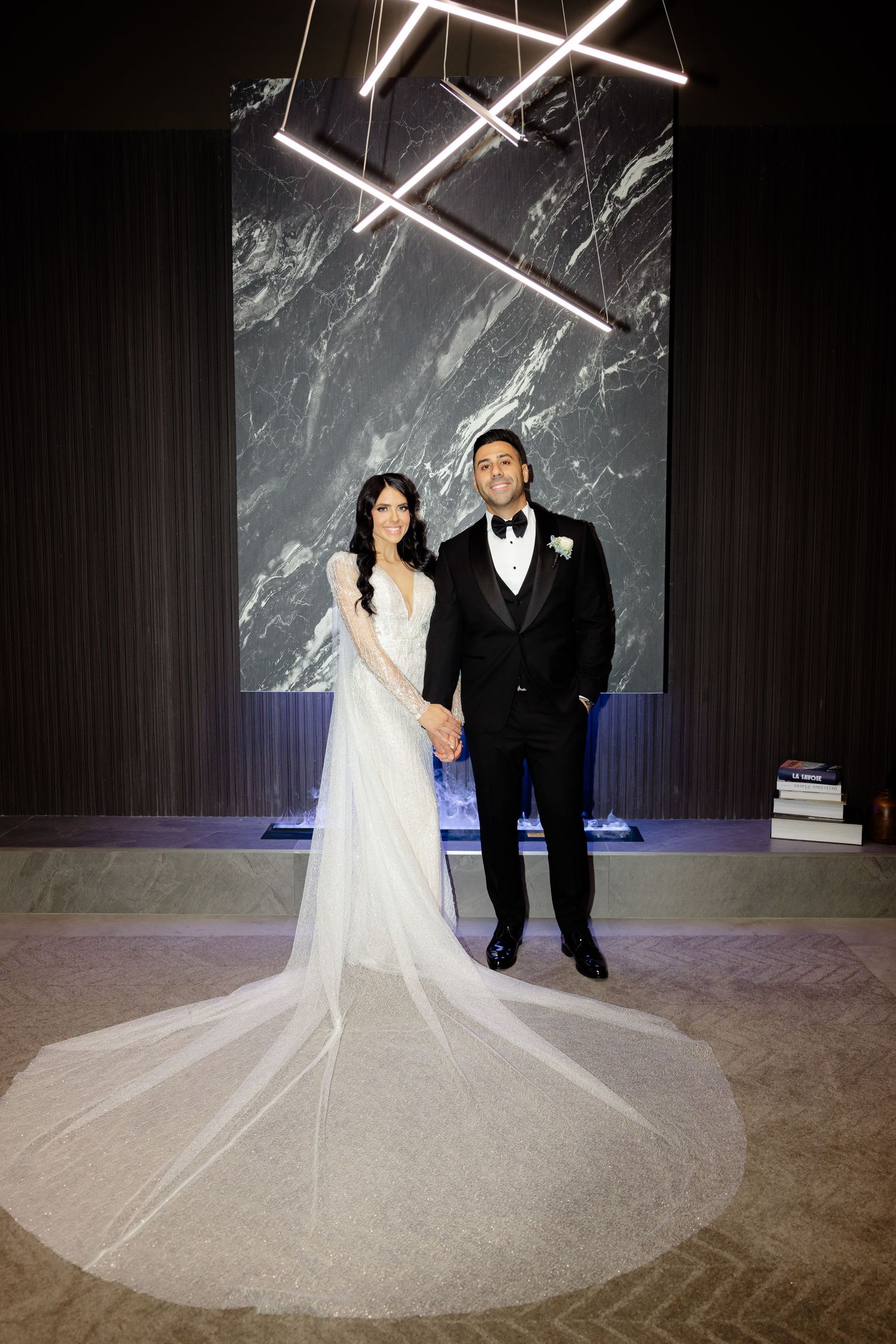 Wedding couple poses formally against marble wall with dramatic cathedral length veil.