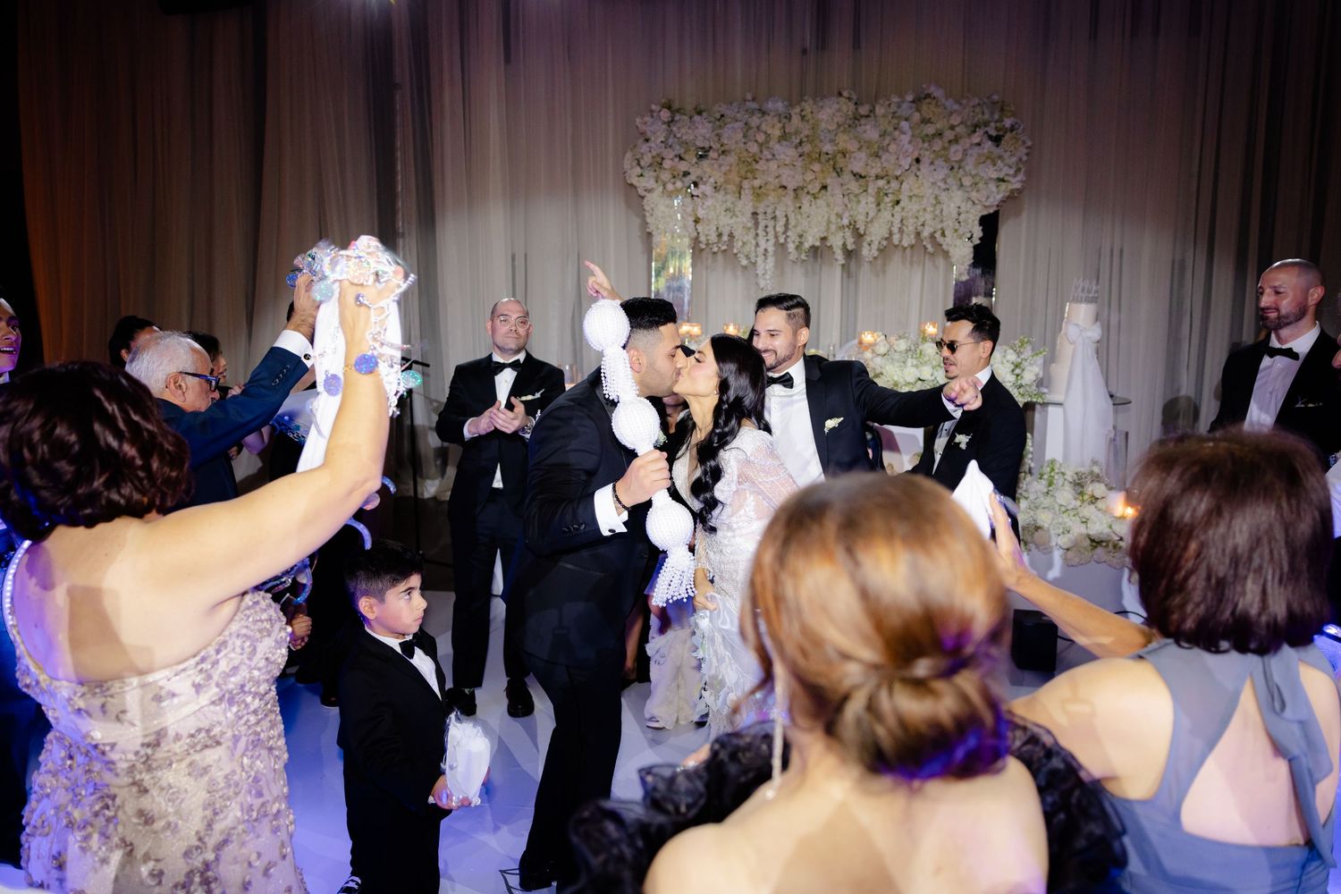 Wedding guests celebrate by raising champagne glasses during a reception under white floral decorations.