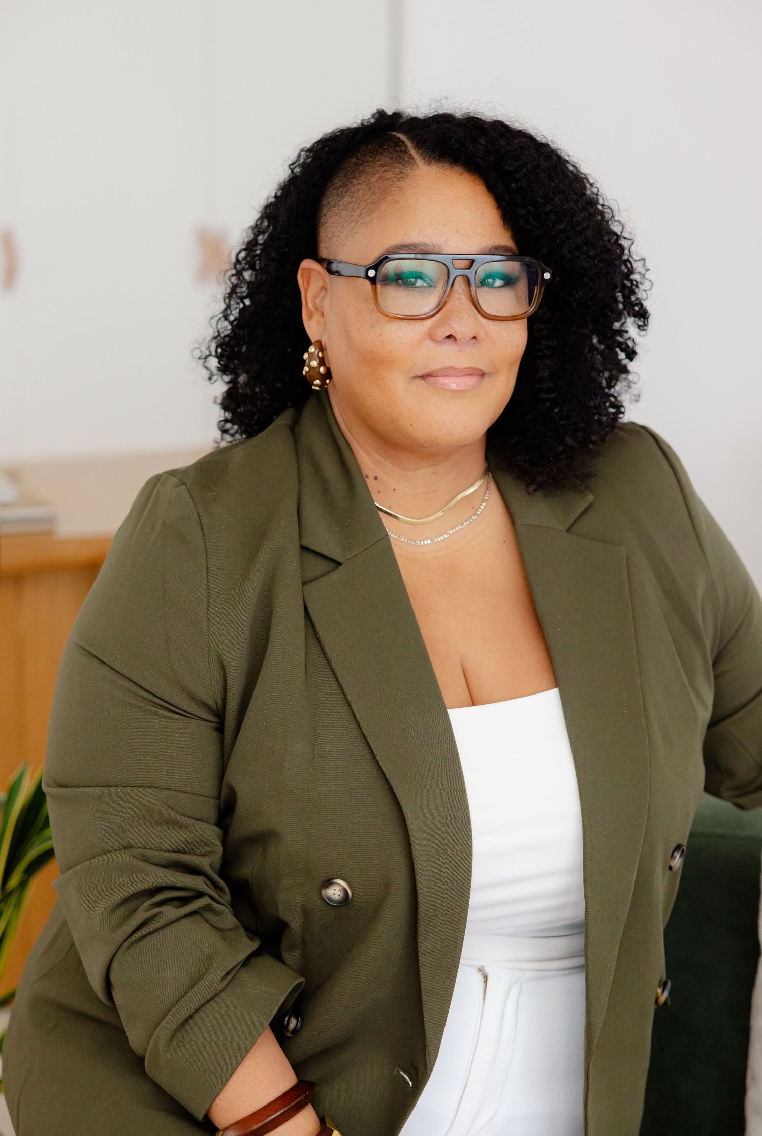 Professional portrait of someone wearing blue glasses, a white top, olive green blazer and gold jewelry against a light backdrop.