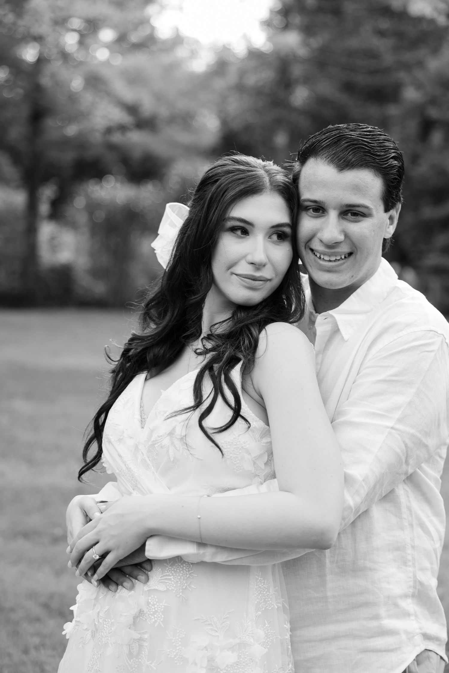 A romantic black and white portrait of a couple embracing outdoors, with her in a white dress and him in a white shirt.