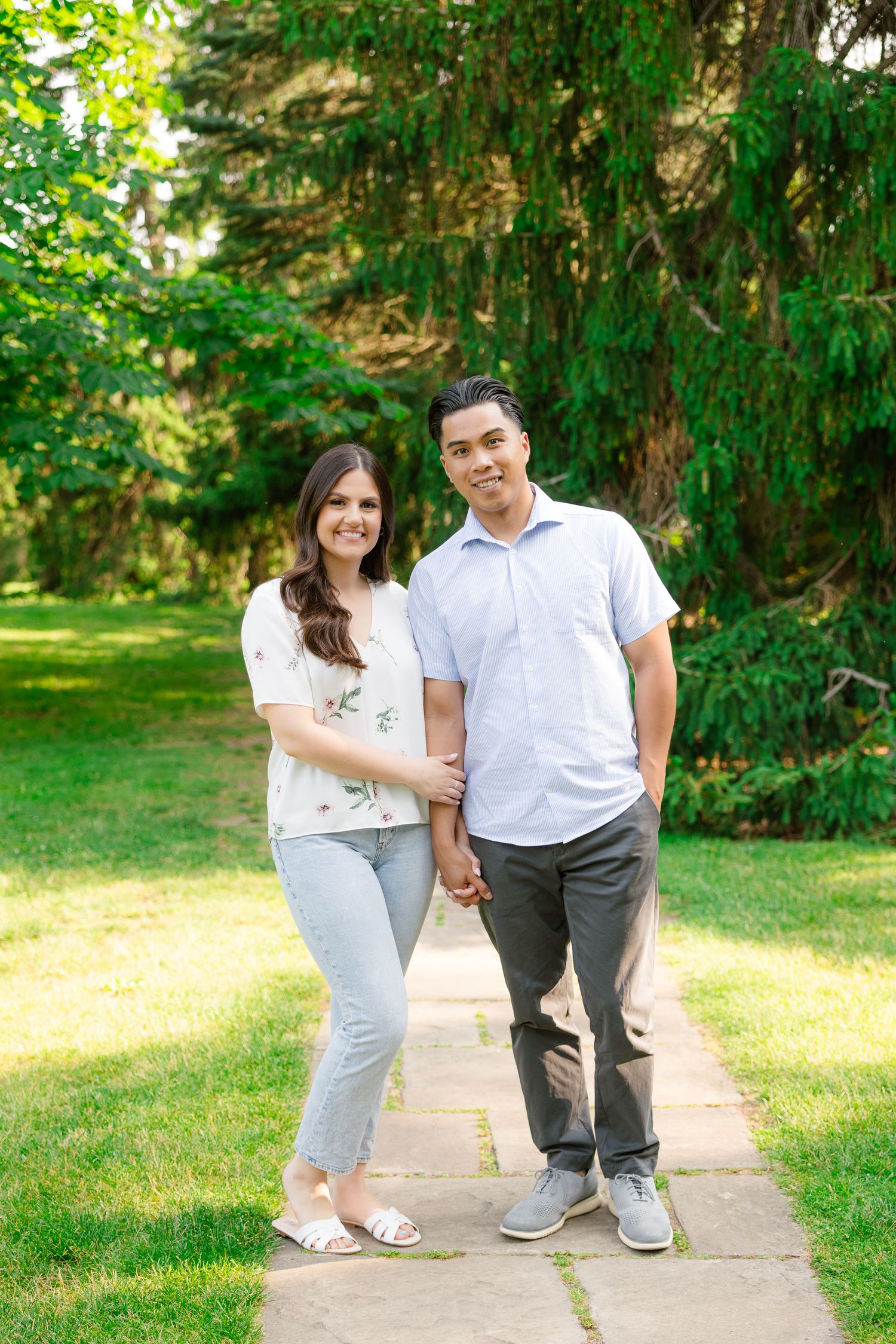 A couple holds hands while standing on a garden path surrounded by lush evergreen trees on a sunny day.