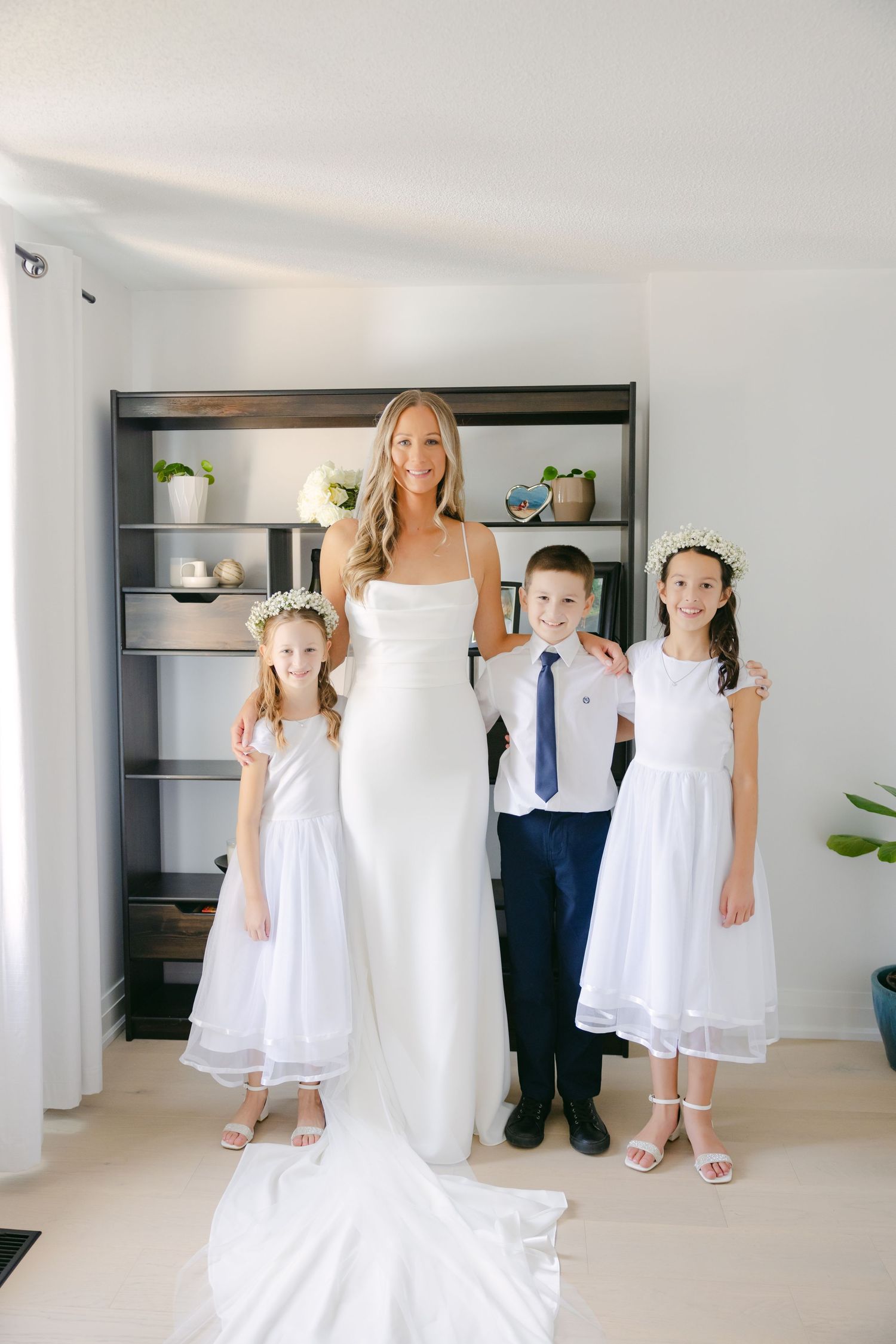 Wedding party members pose in white dresses and formal attire against modern shelving.