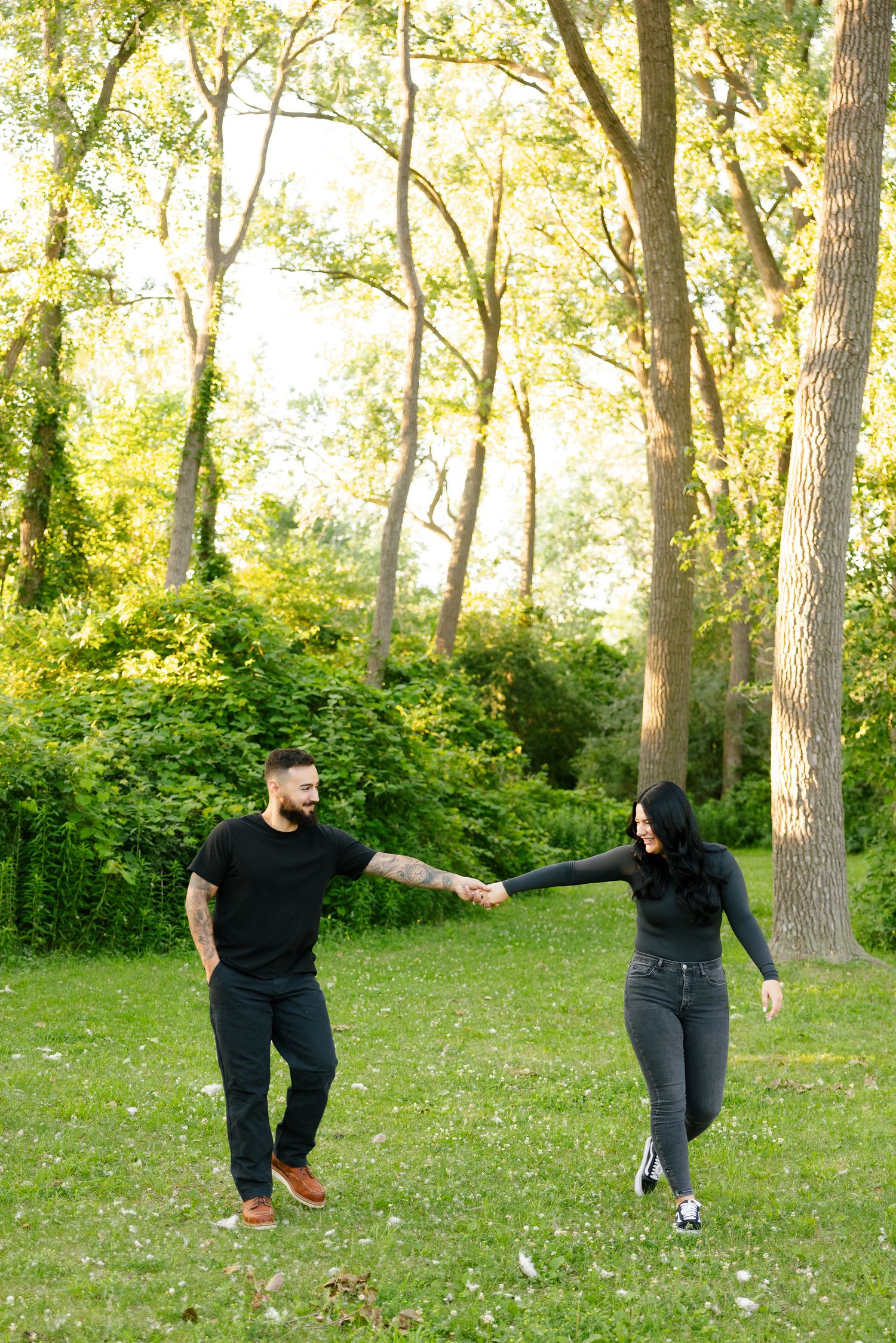 A playful couple holds hands while dancing in a sunny park setting.