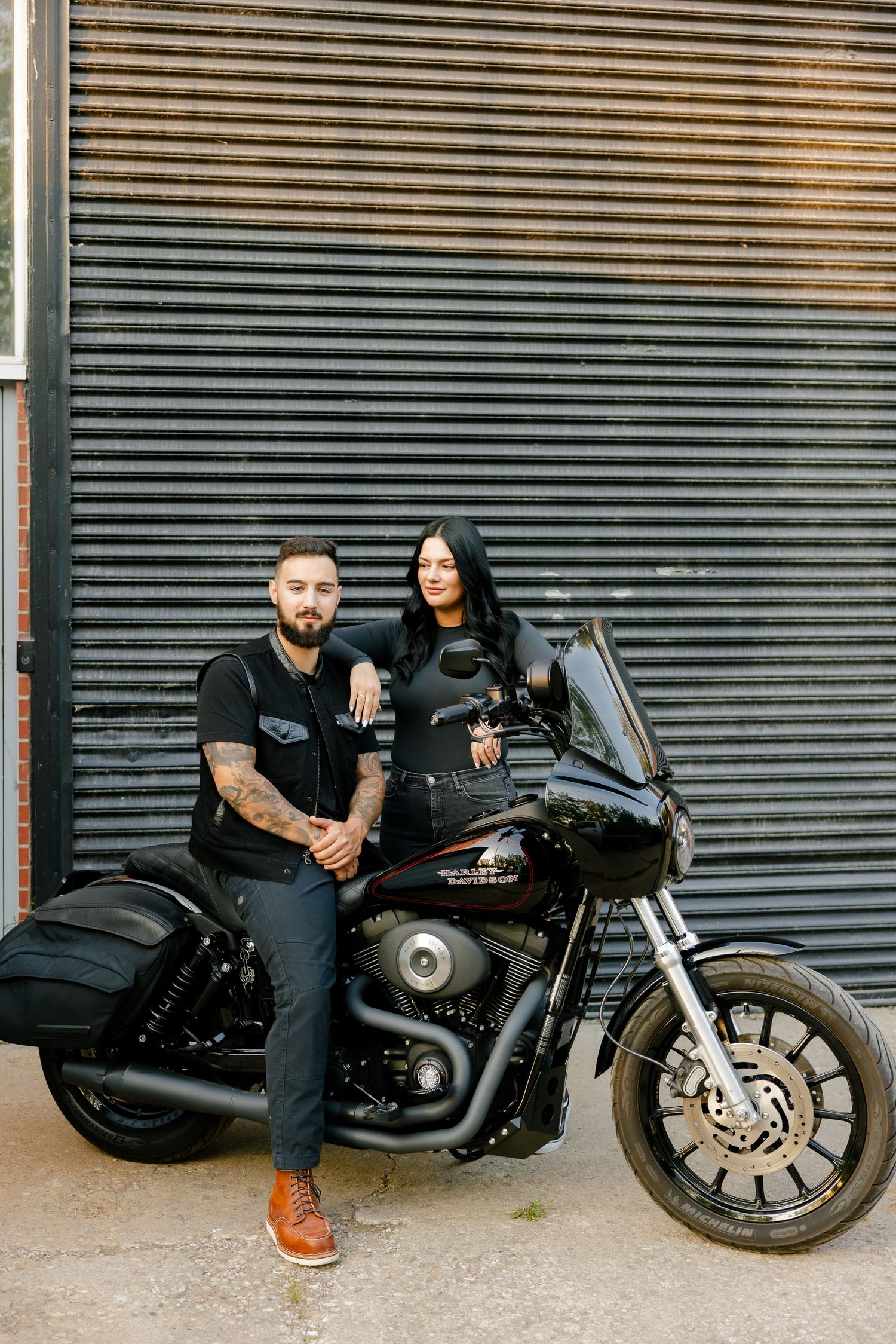 A stylish couple stands with their Harley Davidson motorcycle against an industrial backdrop.