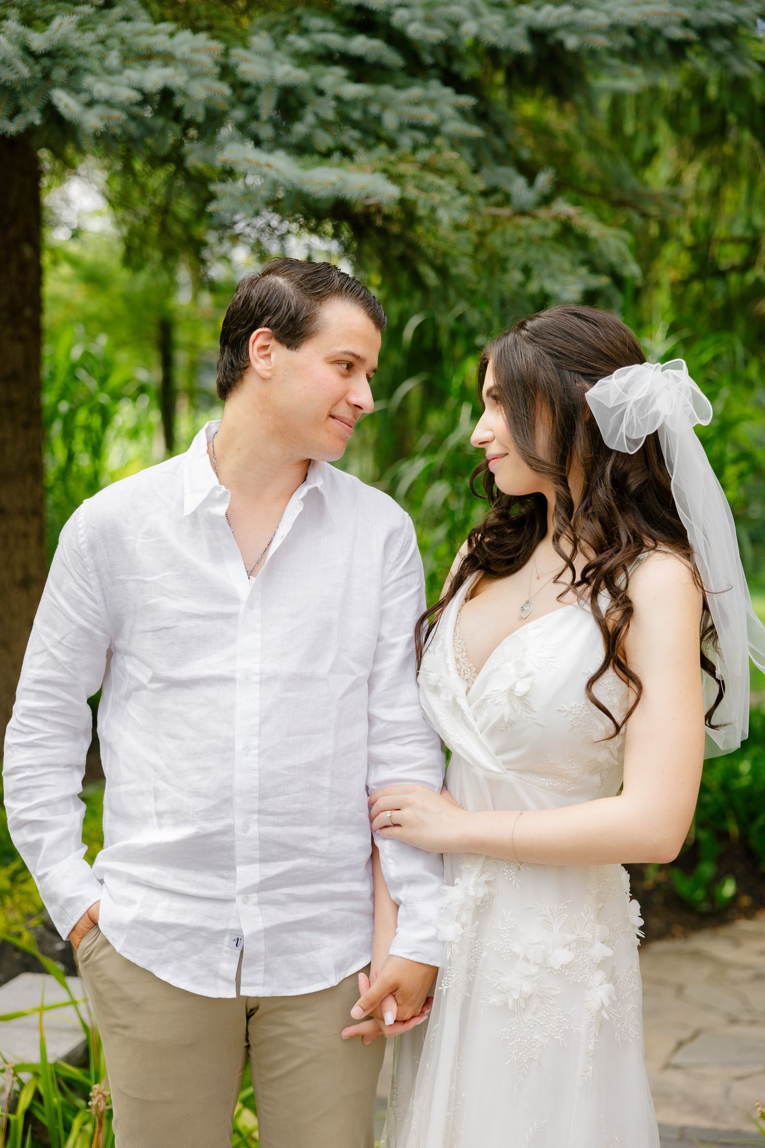 A romantic outdoor wedding portrait of a couple in white attire standing together against a lush garden backdrop.