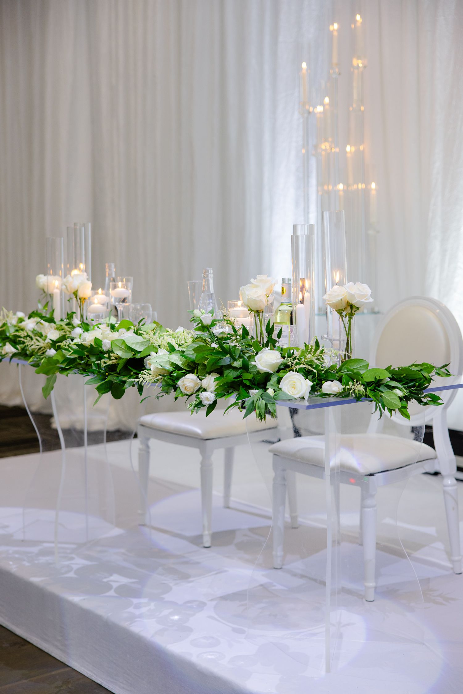 Modern white wedding decor featuring elevated floral arrangements and tall candles on white pedestals.