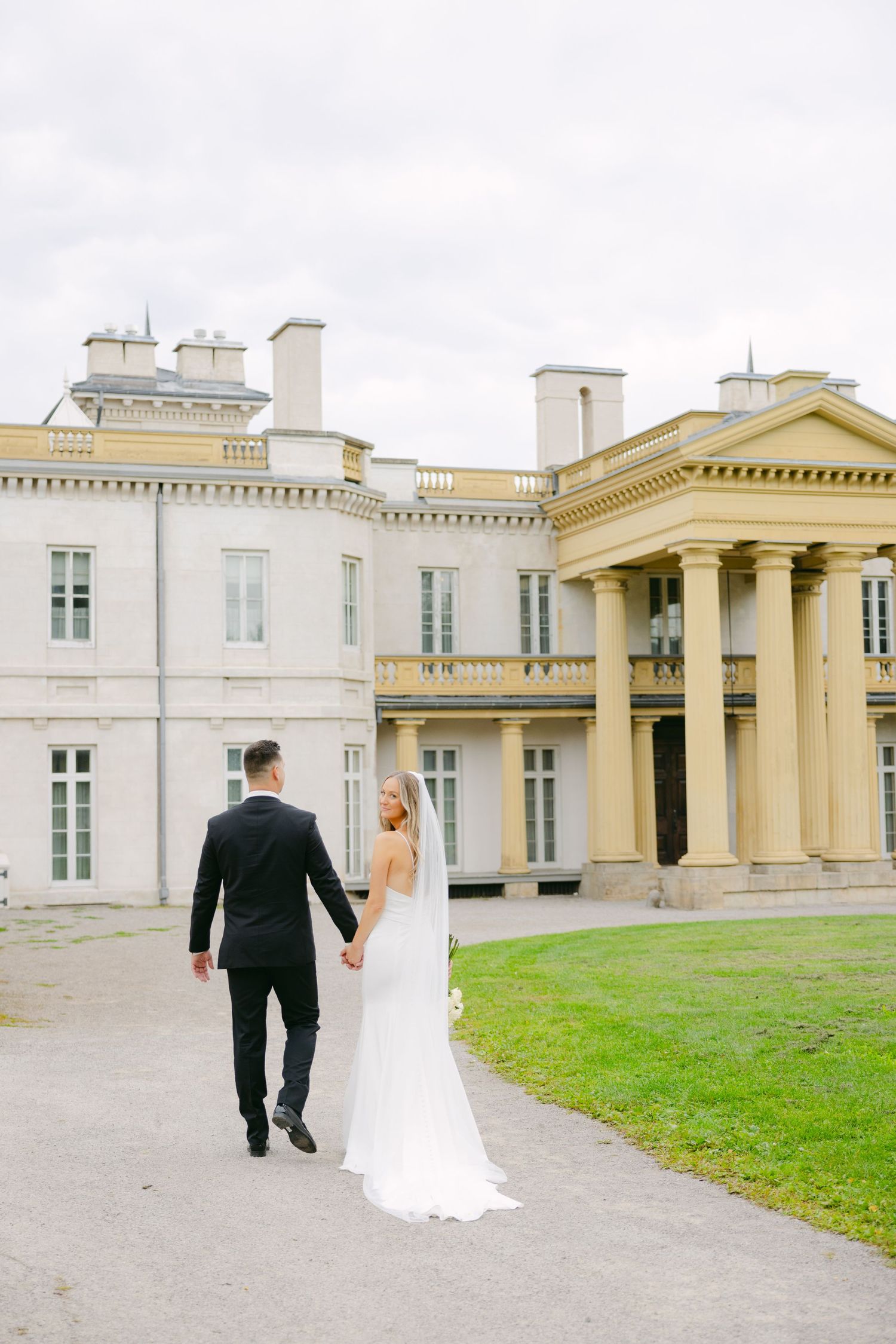 Couple holds hands walking near grand historic mansion with columned entrance.
