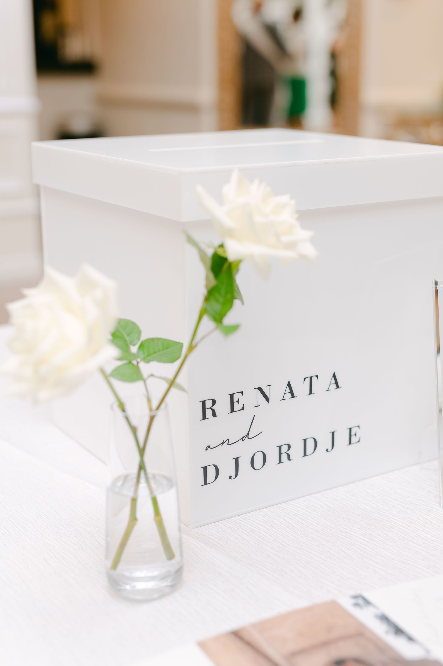 White wedding card box with couple's names and white roses in a small glass vase.
