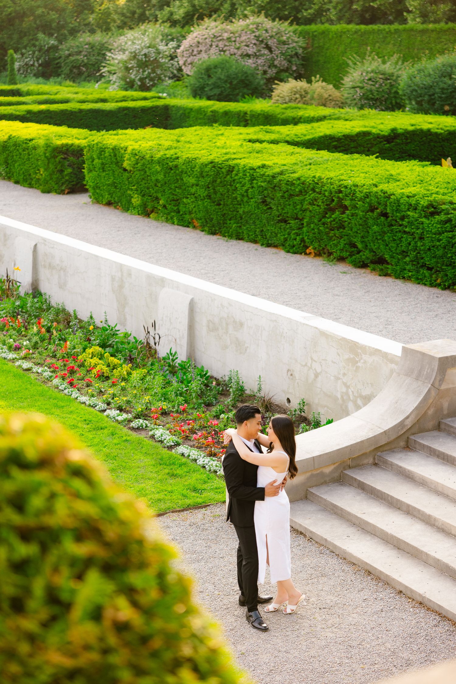 A tender embrace shared at the base of curved stone steps in a lush garden with perfectly maintained hedges.