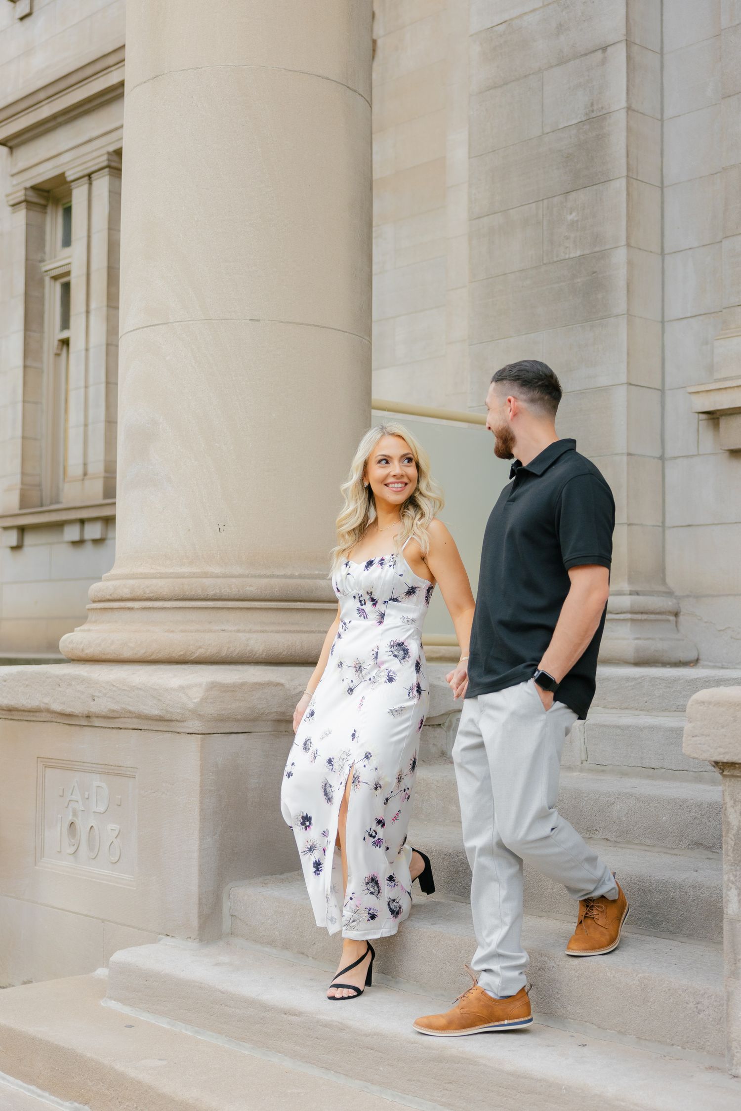 A couple poses on stone steps of a grand building, with her in a floral dress and him in a black polo and khakis.