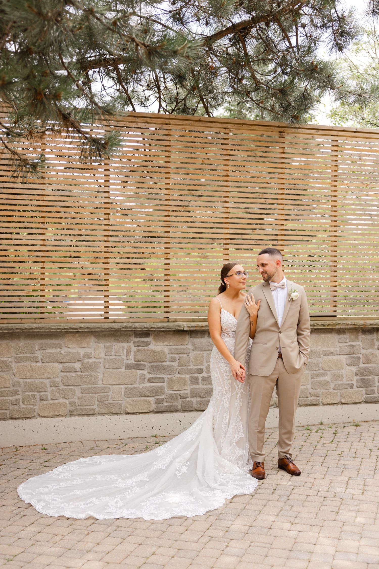 Wedding portrait with long lace train against wooden slat wall on brick paver patio.