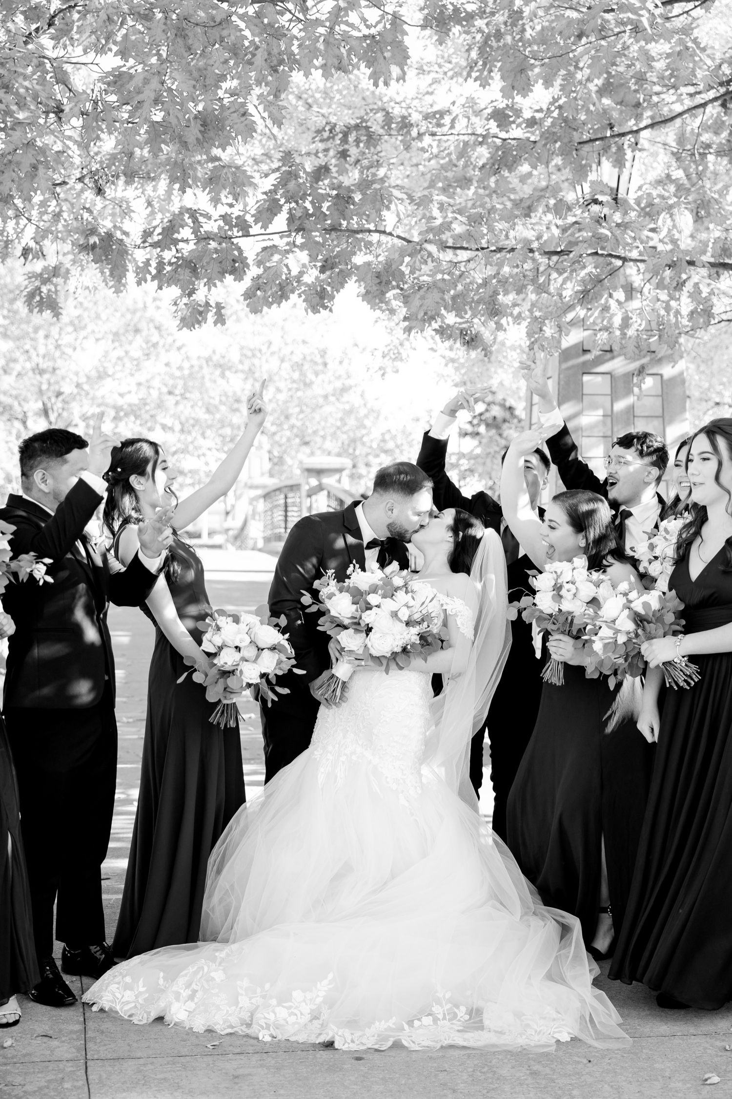 Black and white photo of wedding party celebrating joyfully under tree canopy.
