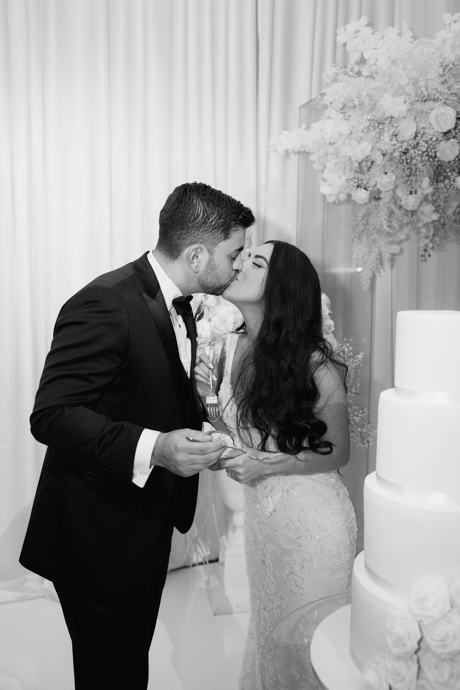 Black and white photo of newlyweds kissing beside their elegant white wedding cake.