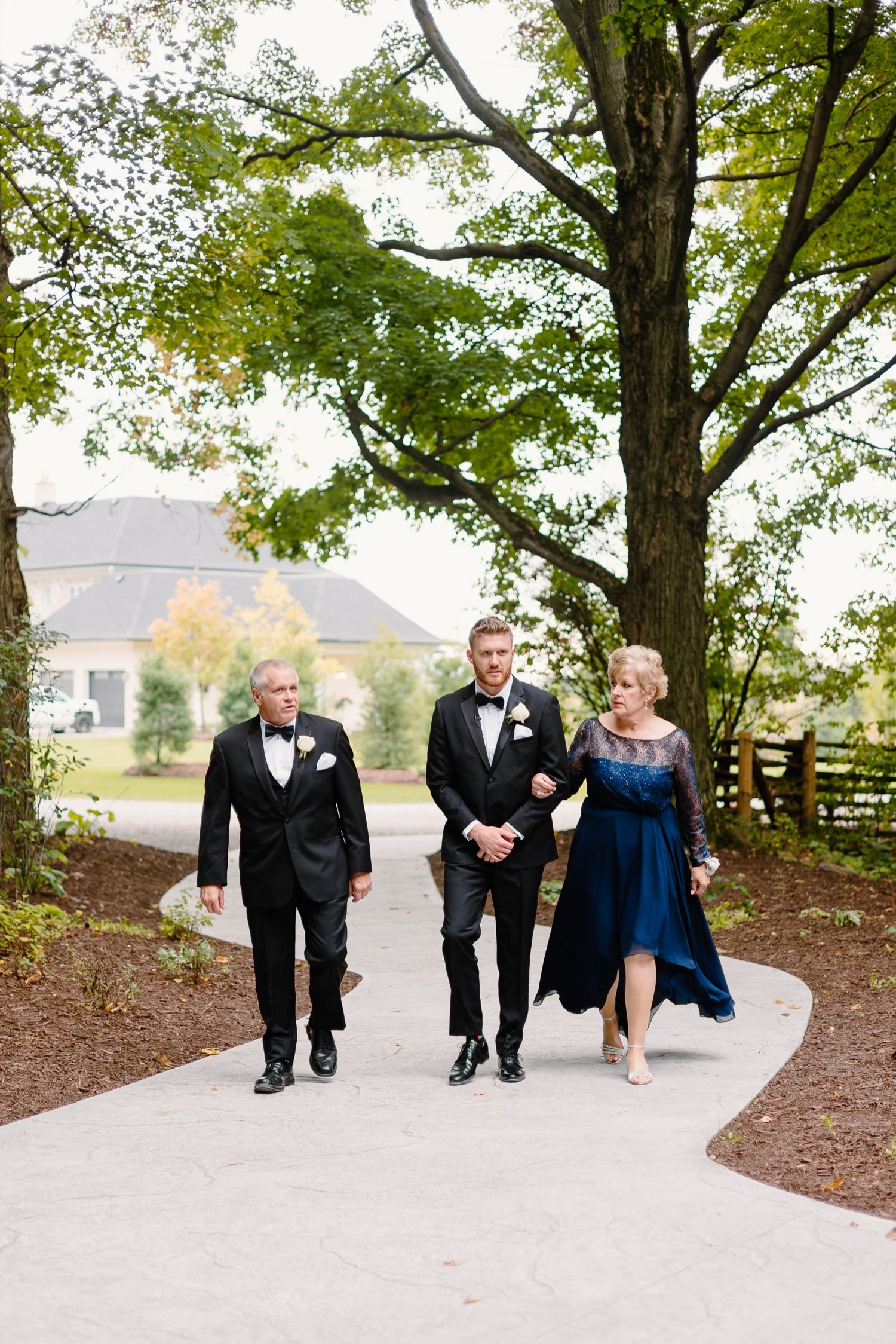 Wedding party members walk along curved path under green canopy of trees.