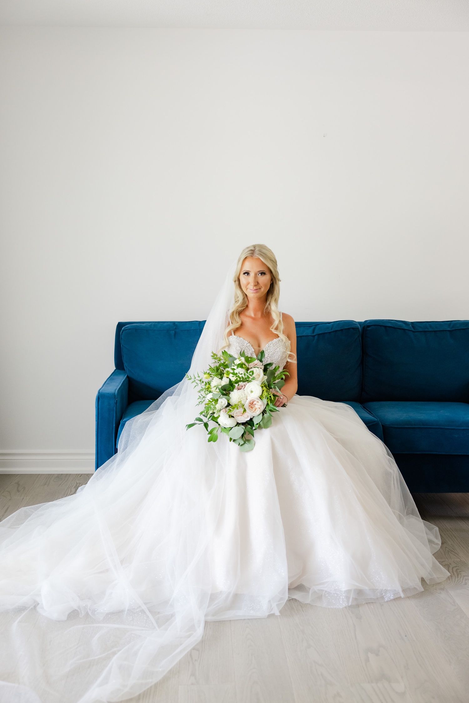 Elegant bride in strapless white dress sits on royal blue sofa holding a cascading bouquet.