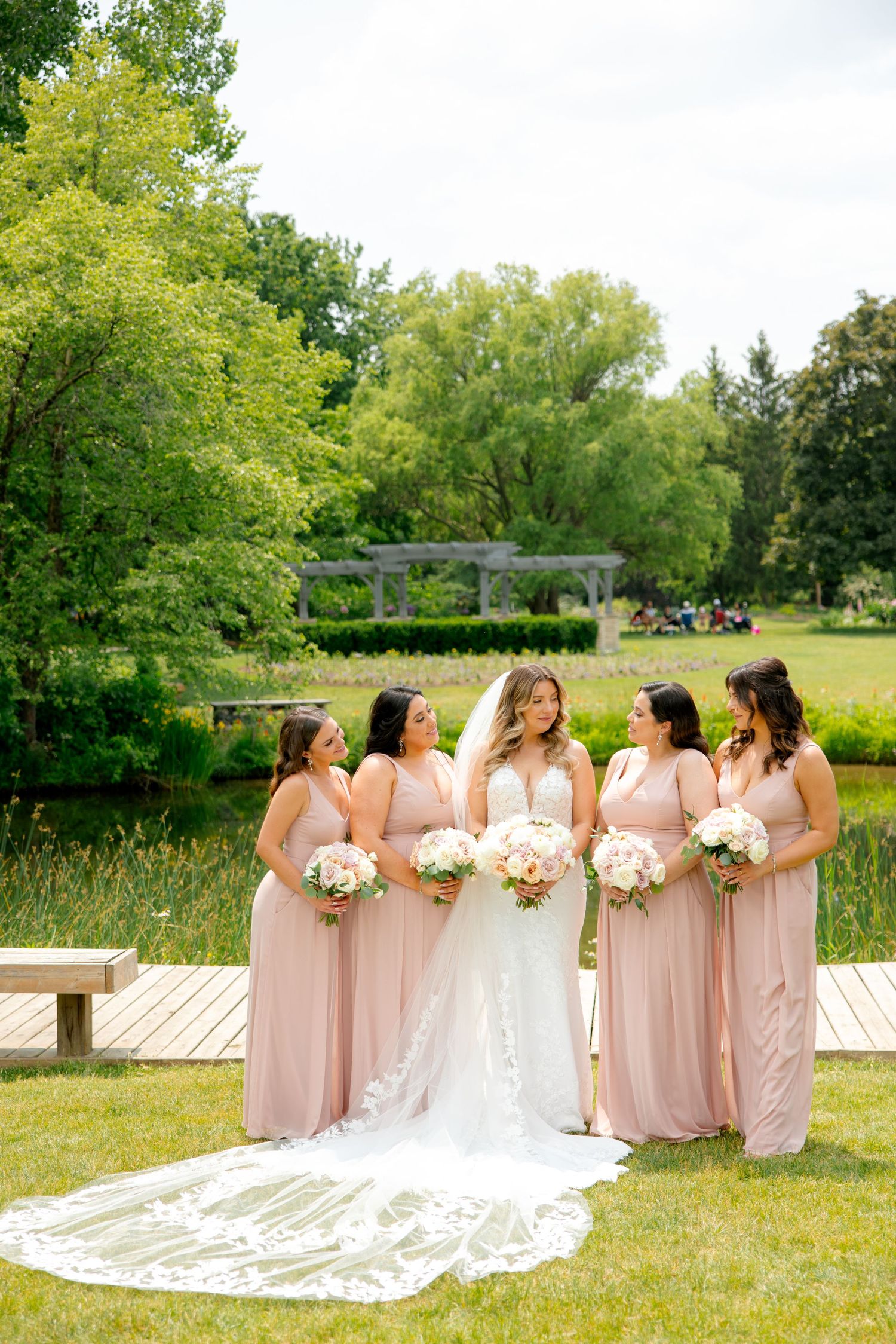 Bridesmaids in soft pink gowns gather around bride with long lace veil on wooden walkway in garden.