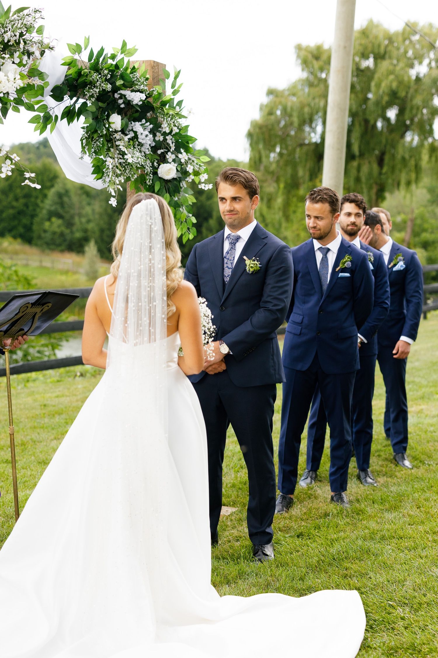 Wedding party stands in a line outdoors wearing navy suits under a floral arch.