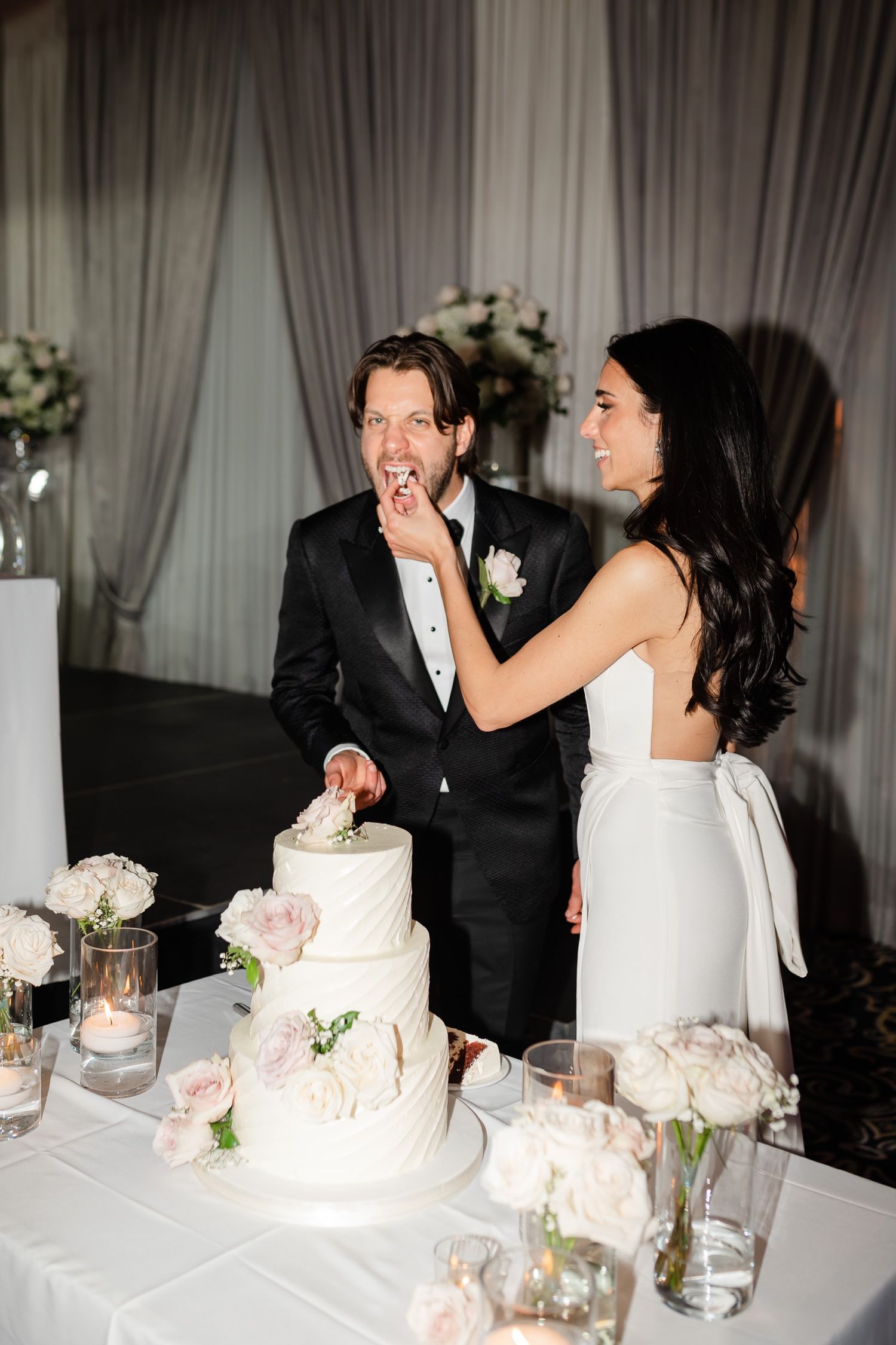 Bride feeds groom a piece of their elegant white tiered wedding cake adorned with pink roses.