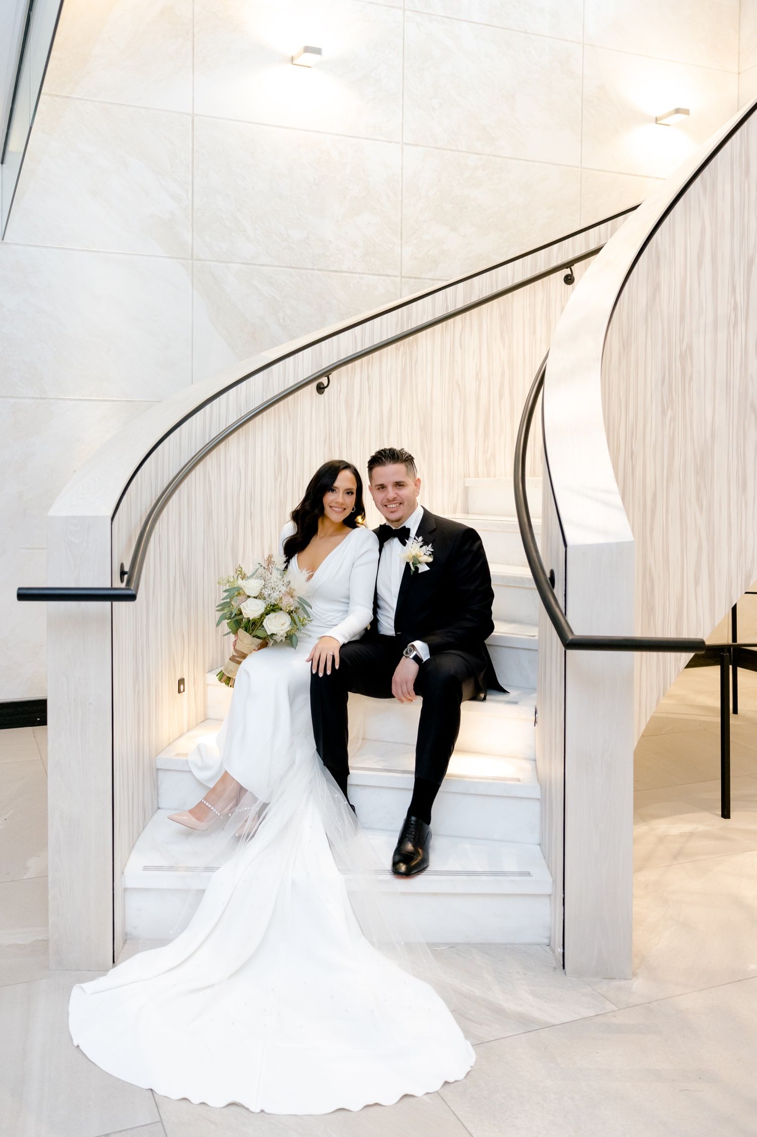 Modern wedding photo of couple seated on elegant white curved staircase with flowing bridal gown train.