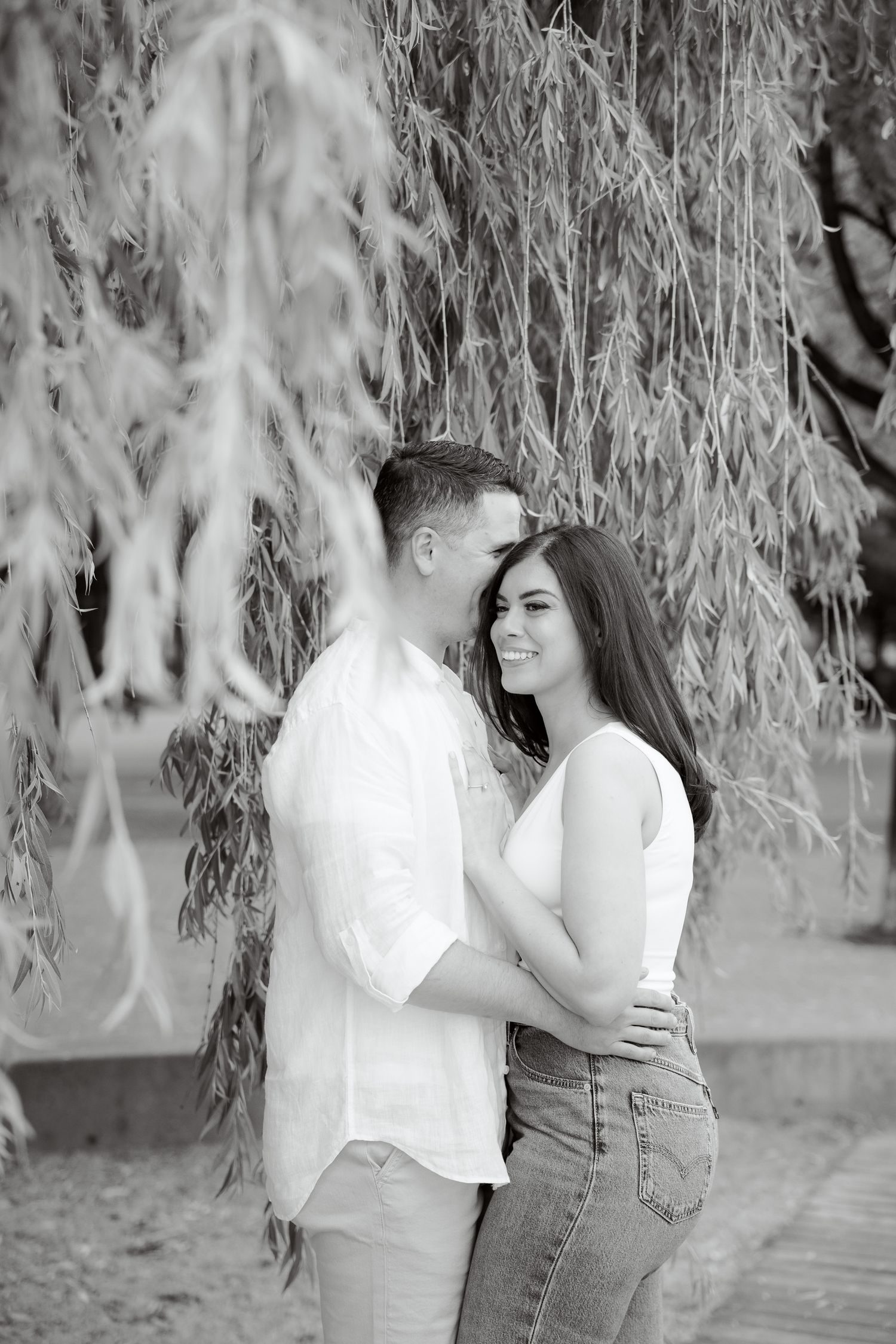A romantic black and white portrait of a couple embracing under drooping willow branches.
