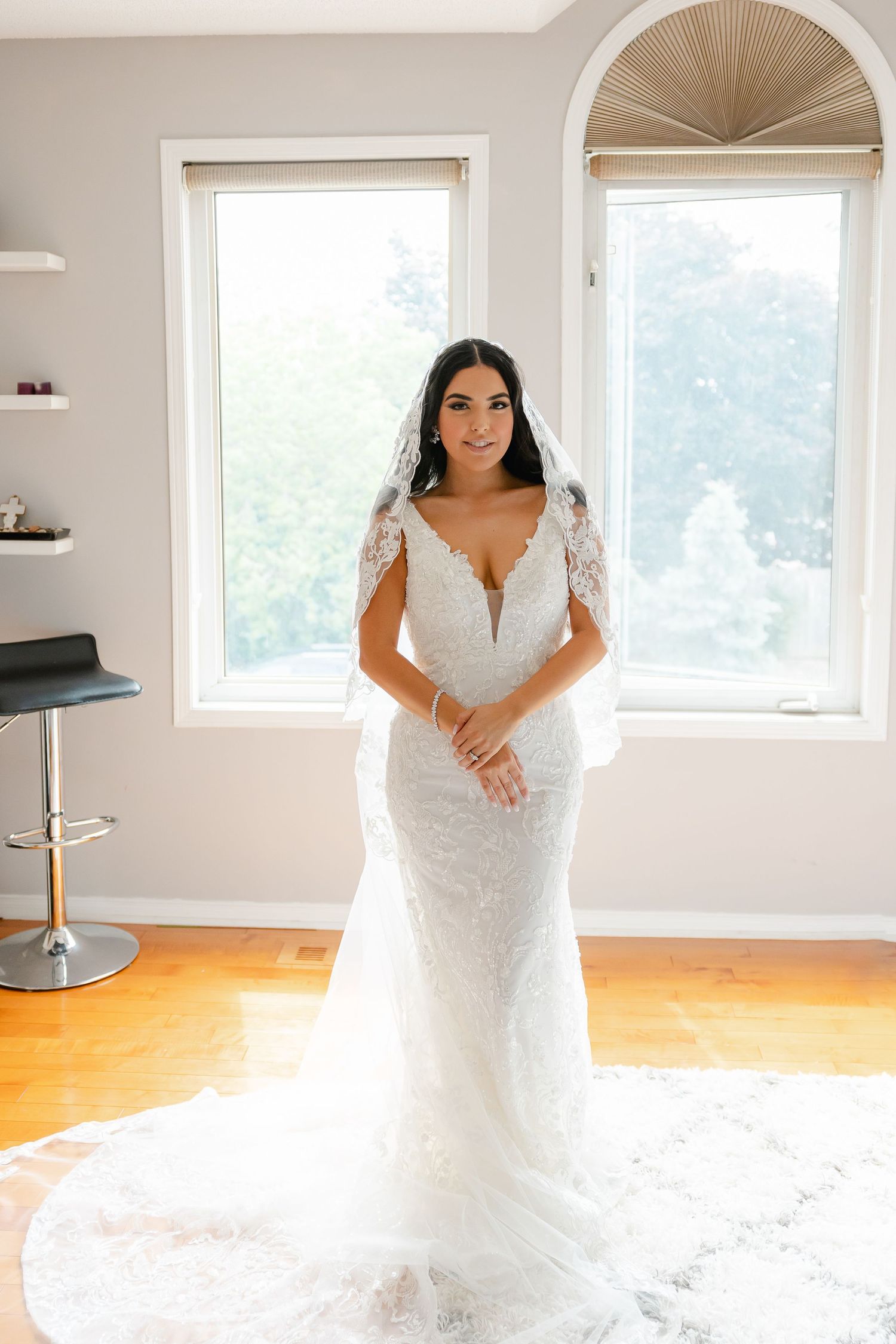 Elegant bride in fitted lace wedding gown poses by sunlit windows.