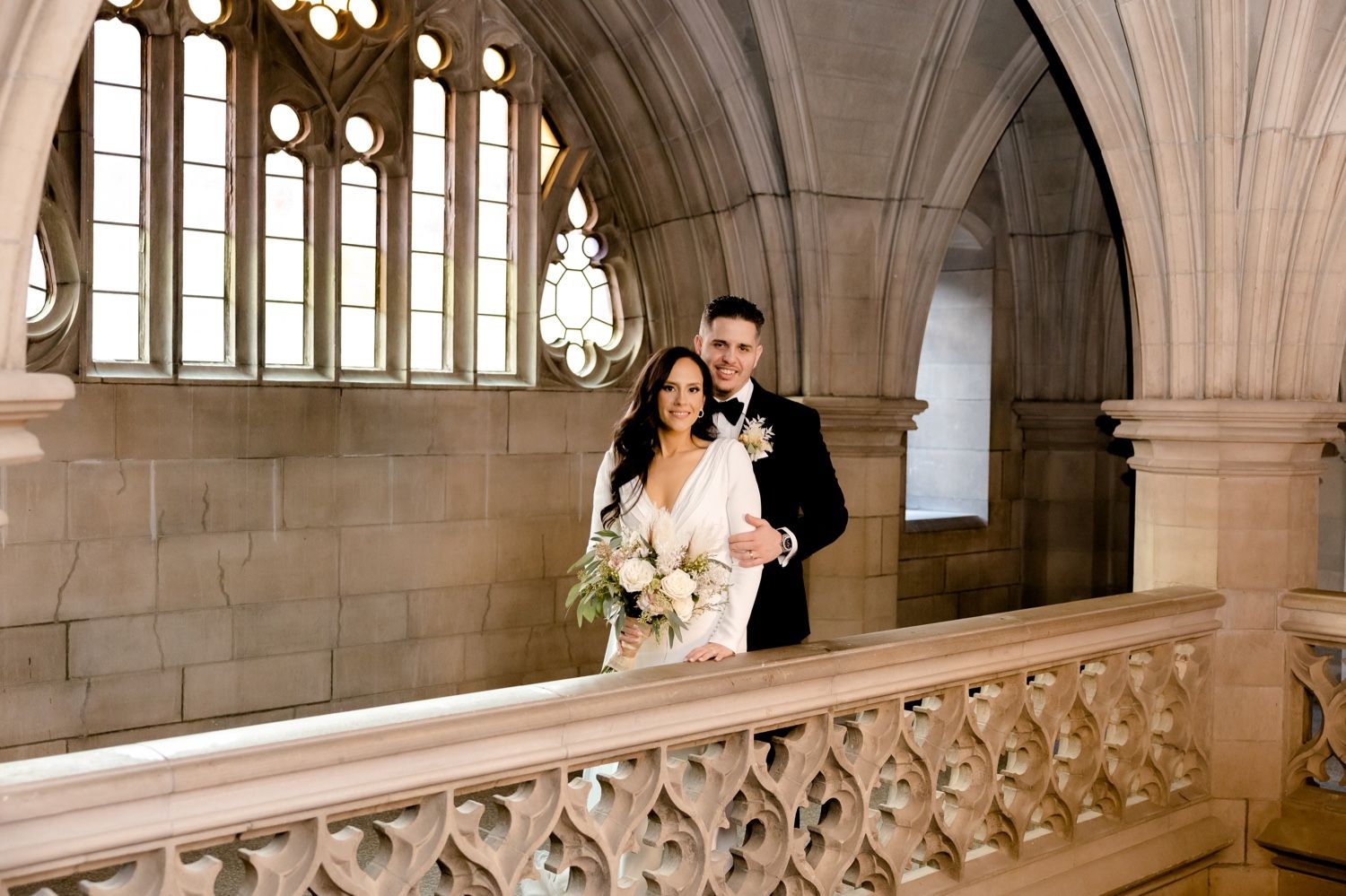 Wedding couple stands by ornate stone railing with Gothic windows and vaulted ceiling.