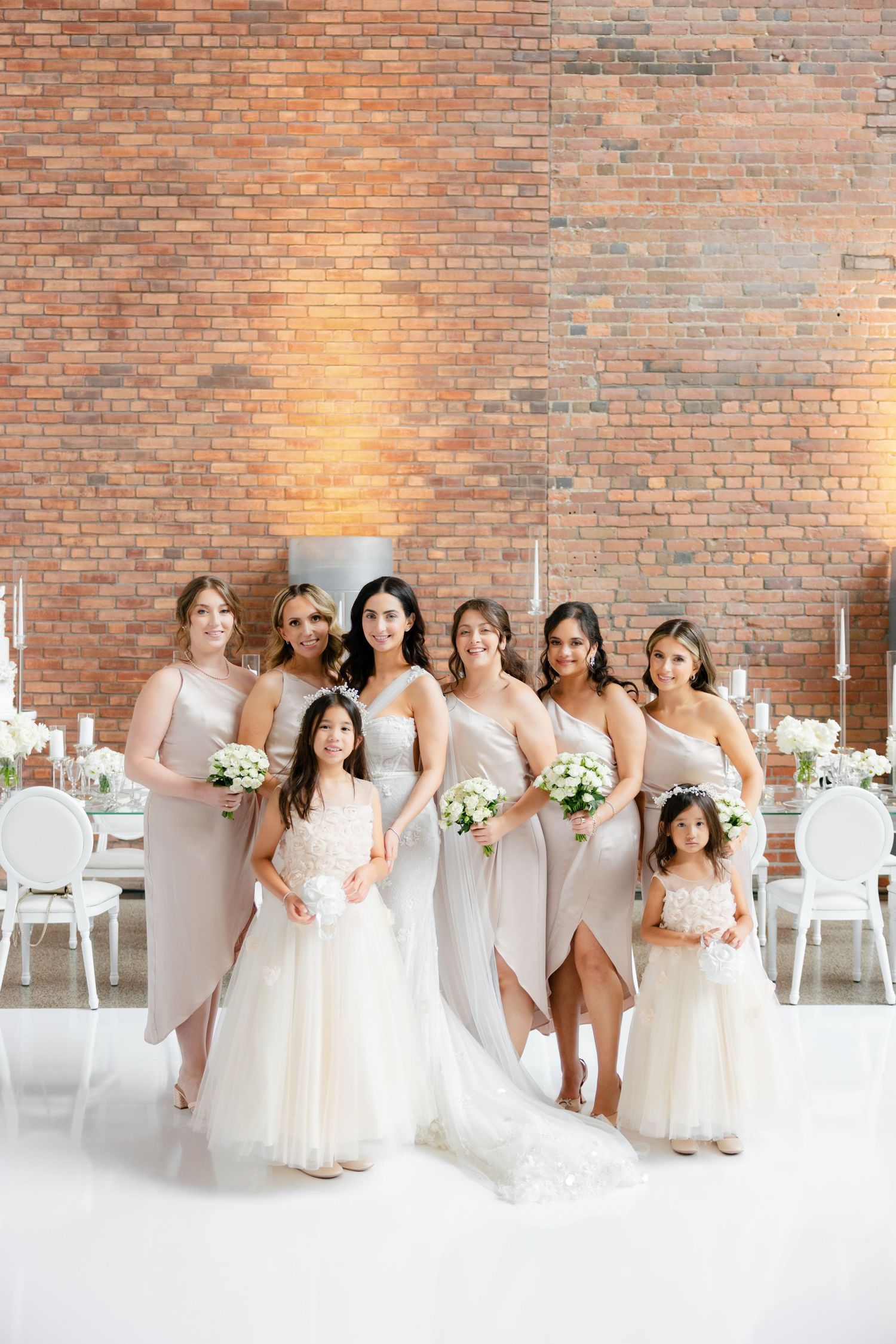 Bridal party in blush dresses poses against brick wall in elegant white reception space.