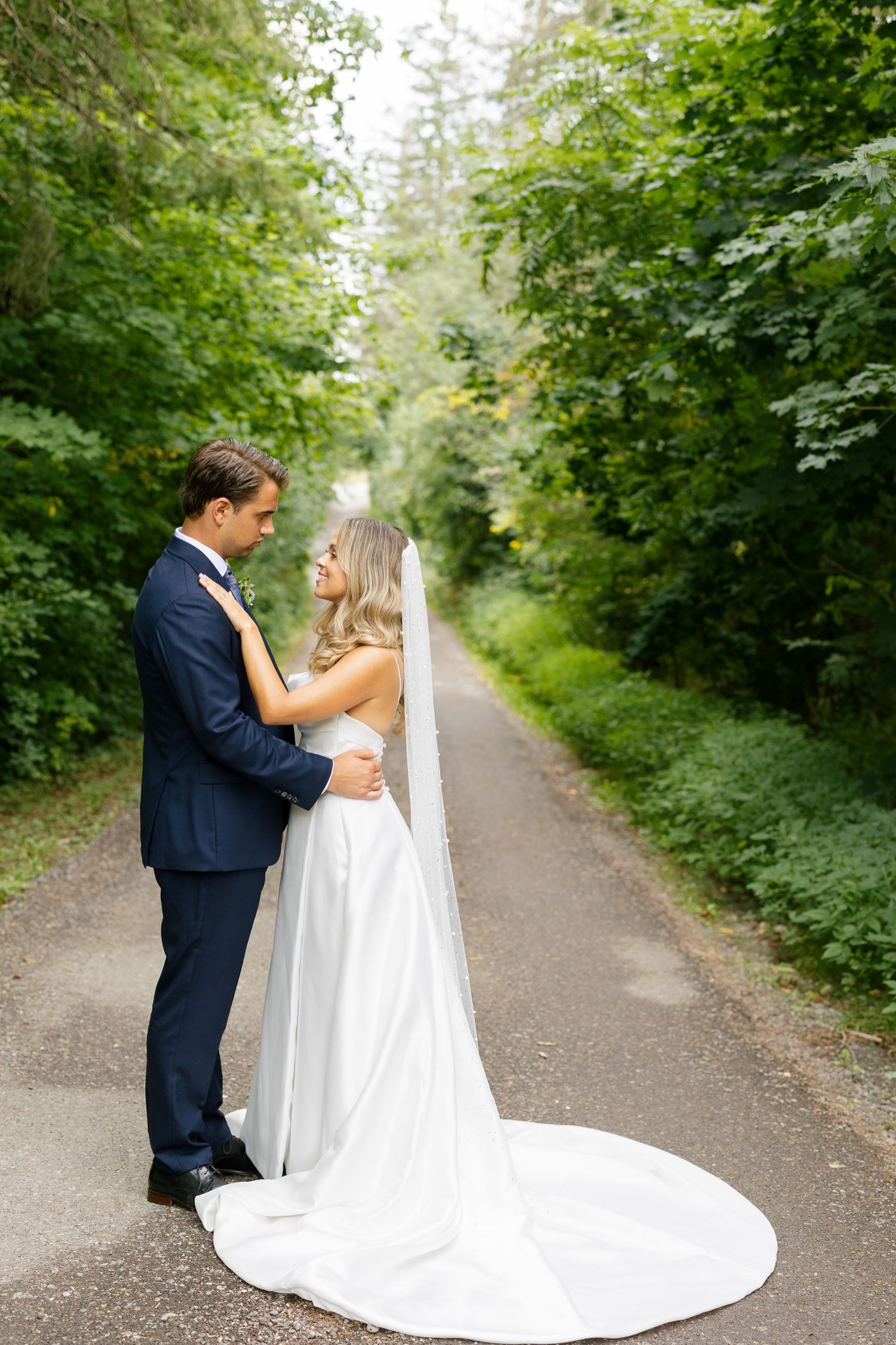 Elegant couple embraces on forest path with flowing white wedding dress train.
