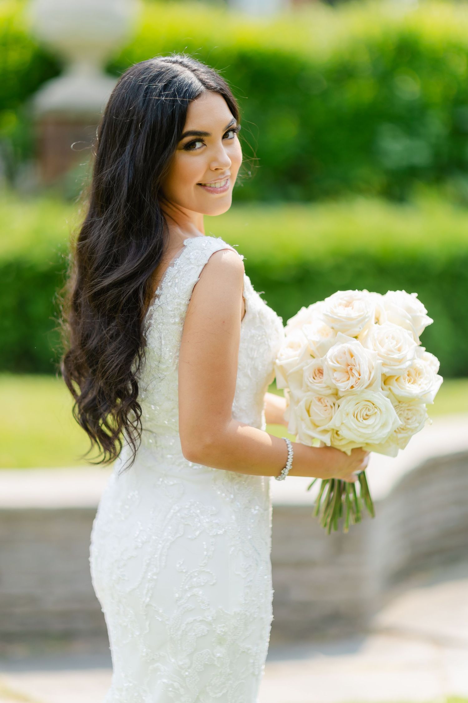 Elegant bride in fitted lace dress holds white rose bouquet while posing in garden setting.