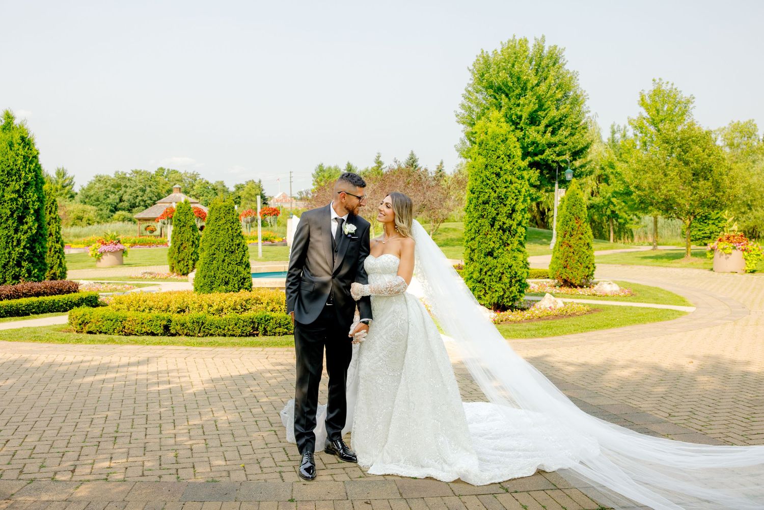 Scenic wedding photo with couple on brick walkway framed by perfectly trimmed evergreens.