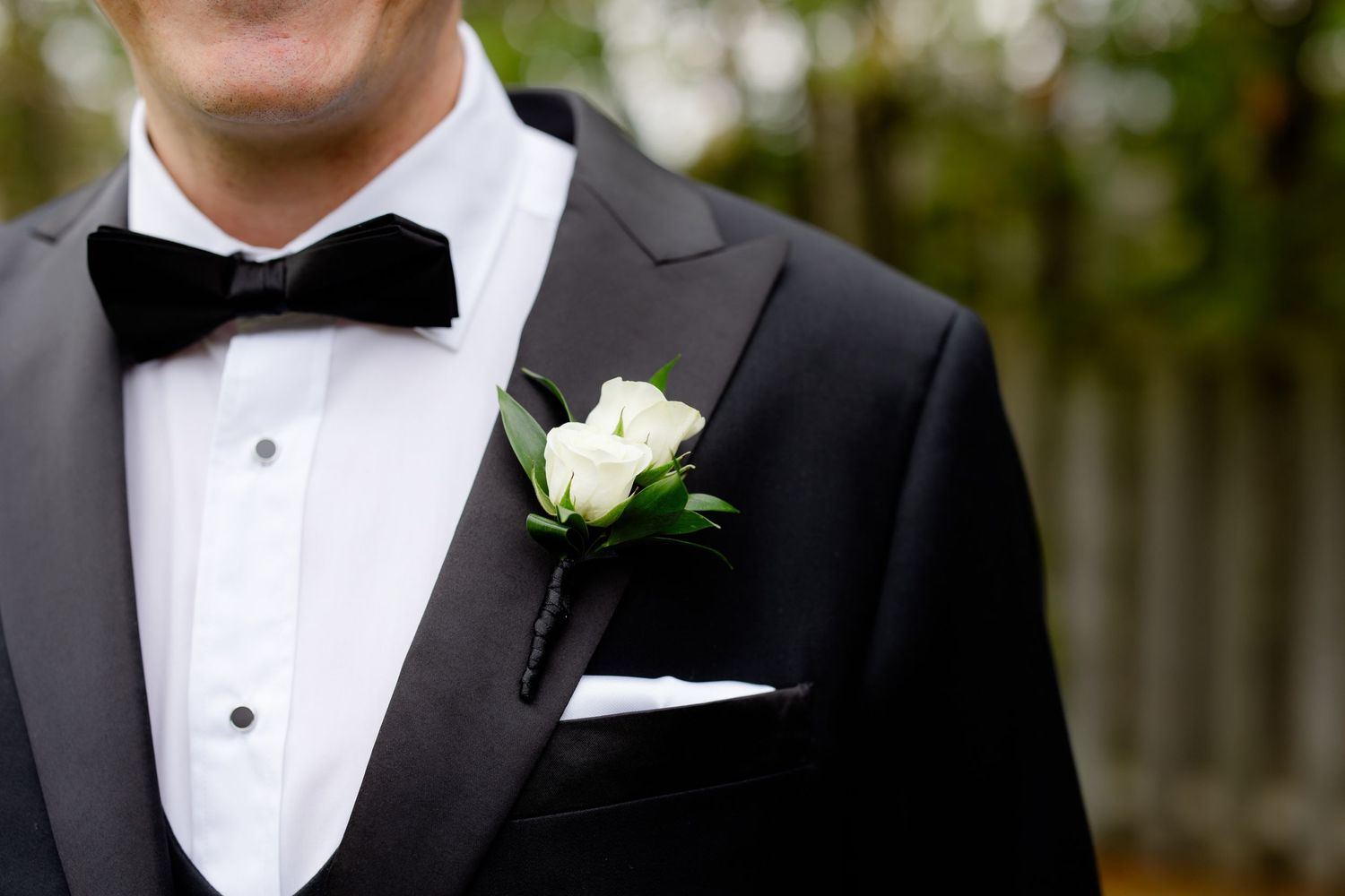 Close-up of a black tuxedo with white boutonniere and bow tie against a blurred background.