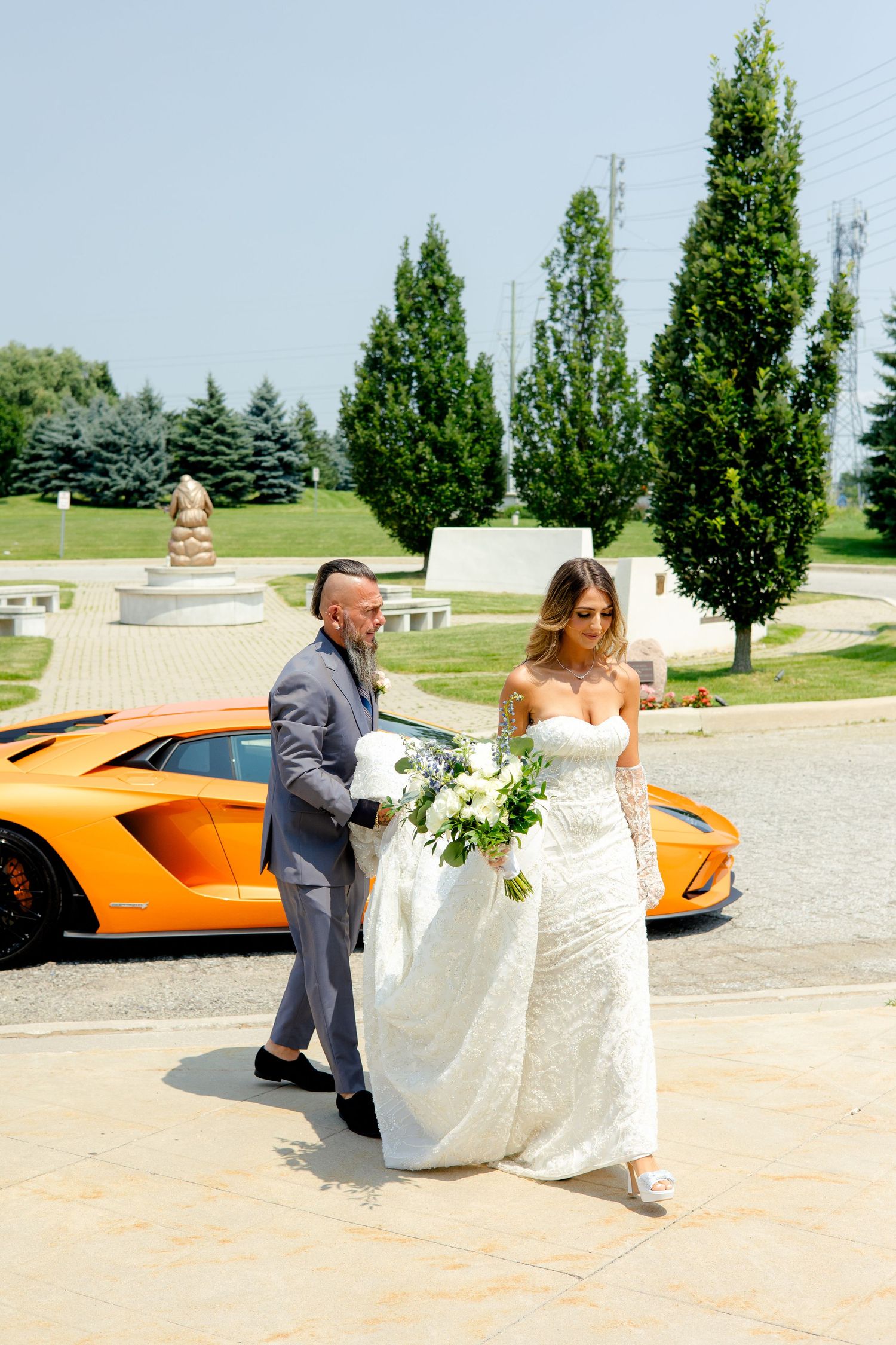 A couple walks together past an orange sports car, with the bride carrying a white floral bouquet.