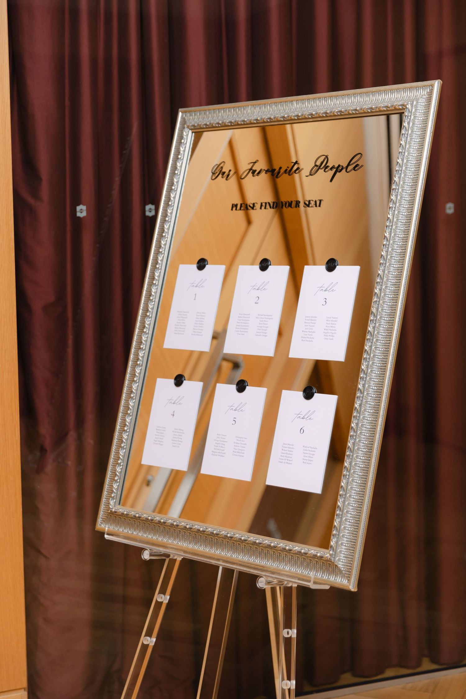 Wedding seating chart displayed in a silver frame on an easel against burgundy curtains.