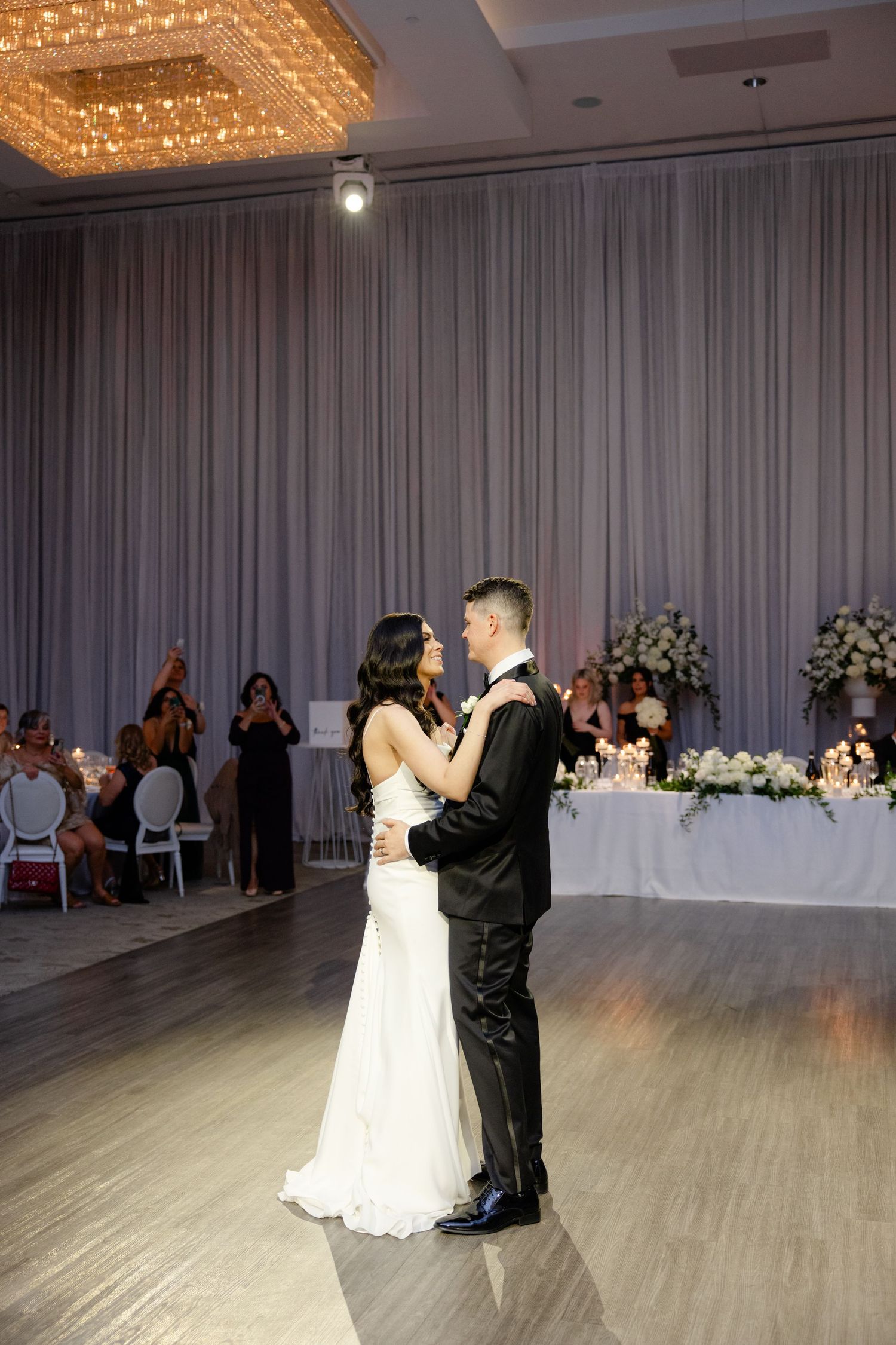Couple dances together in elegant ballroom with crystal chandelier and draped walls.