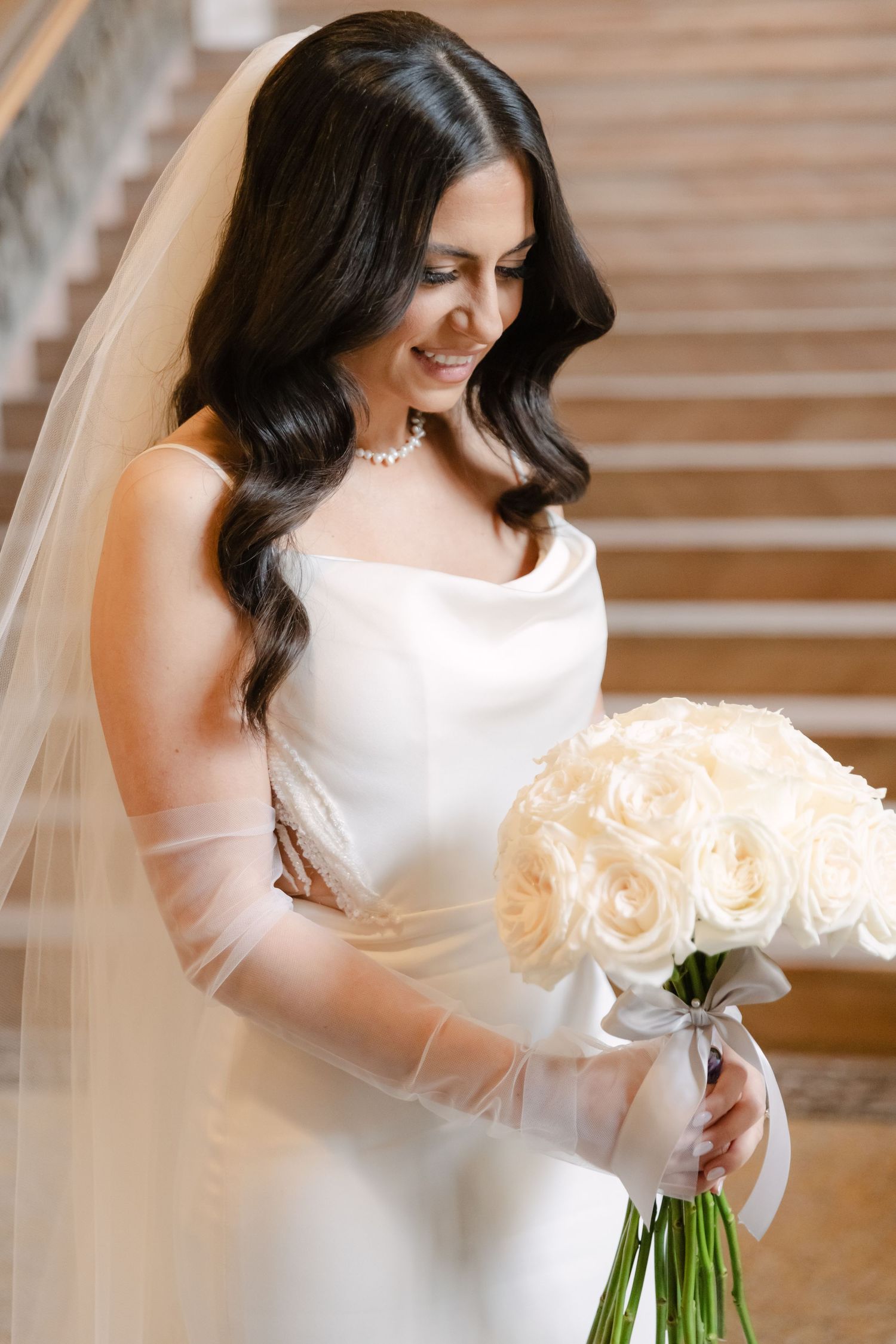 An elegant bride in a white dress holds a bouquet of cream roses against a stone staircase backdrop.