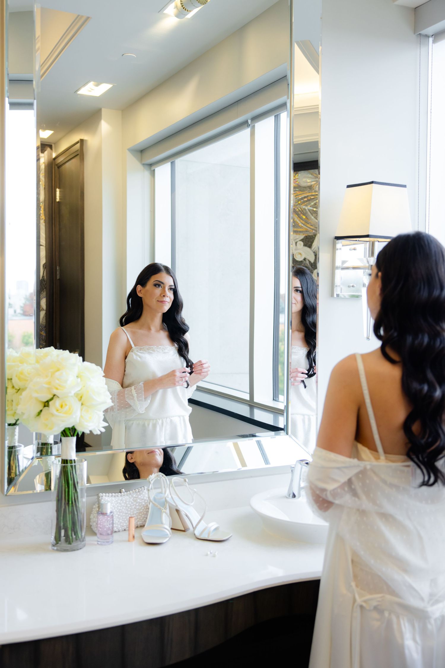 Bride in white getting ready in modern bathroom with mirror reflection and floral bouquet.