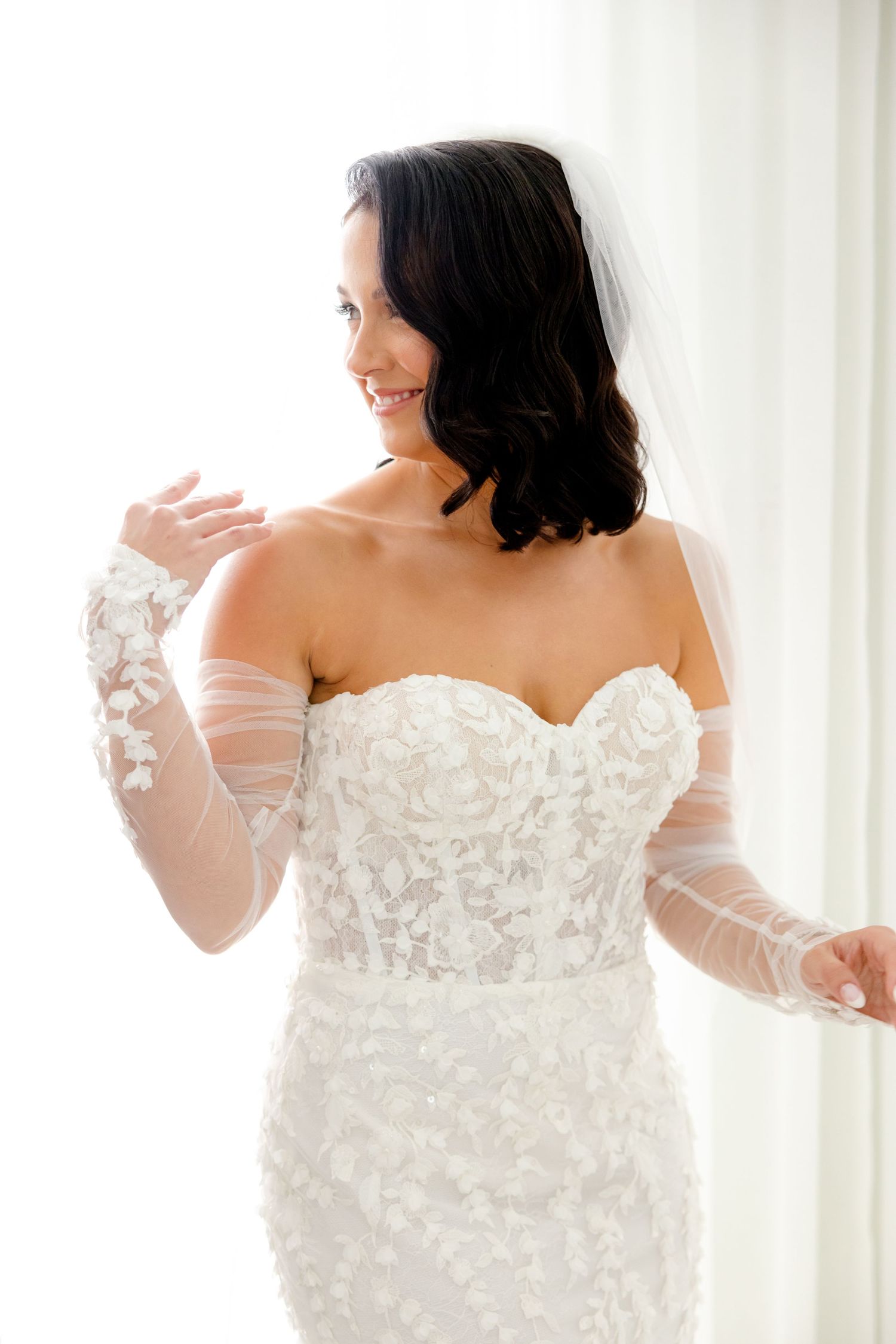 Side view of a bride in an elegant strapless lace wedding dress with sheer sleeves.