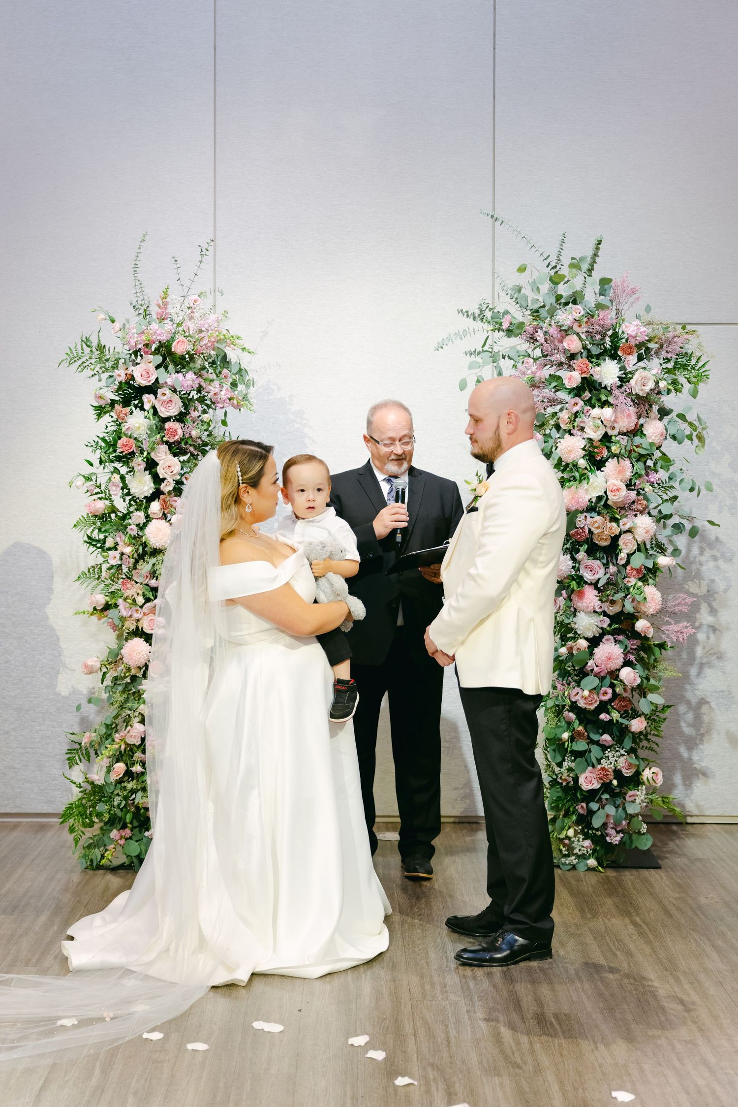 Wedding ceremony under beautiful pink floral arch with officiant and small child.