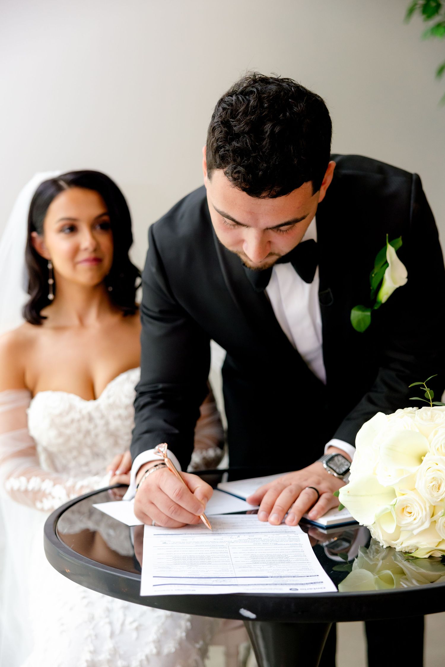 Close-up of groom signing marriage certificate while bride watches in elegant setting.
