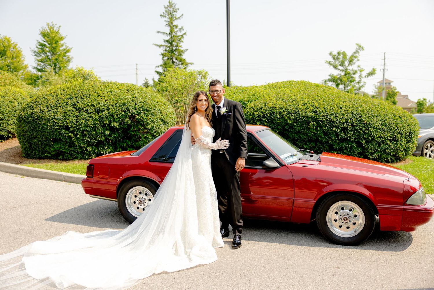 Bride in flowing white gown and groom in black suit pose with vintage red car.