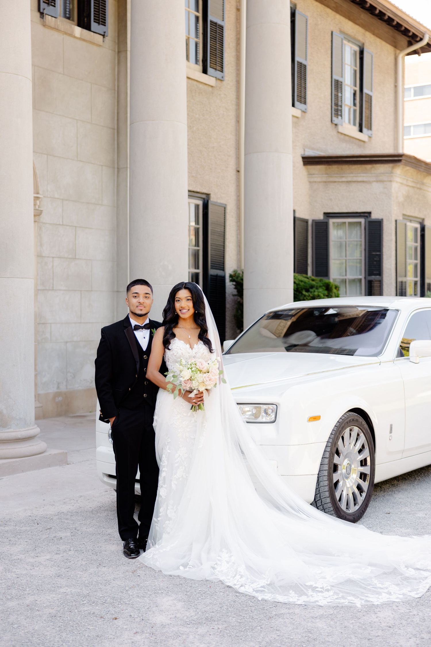Wedding couple stands elegantly next to white luxury car with classical architecture in background.