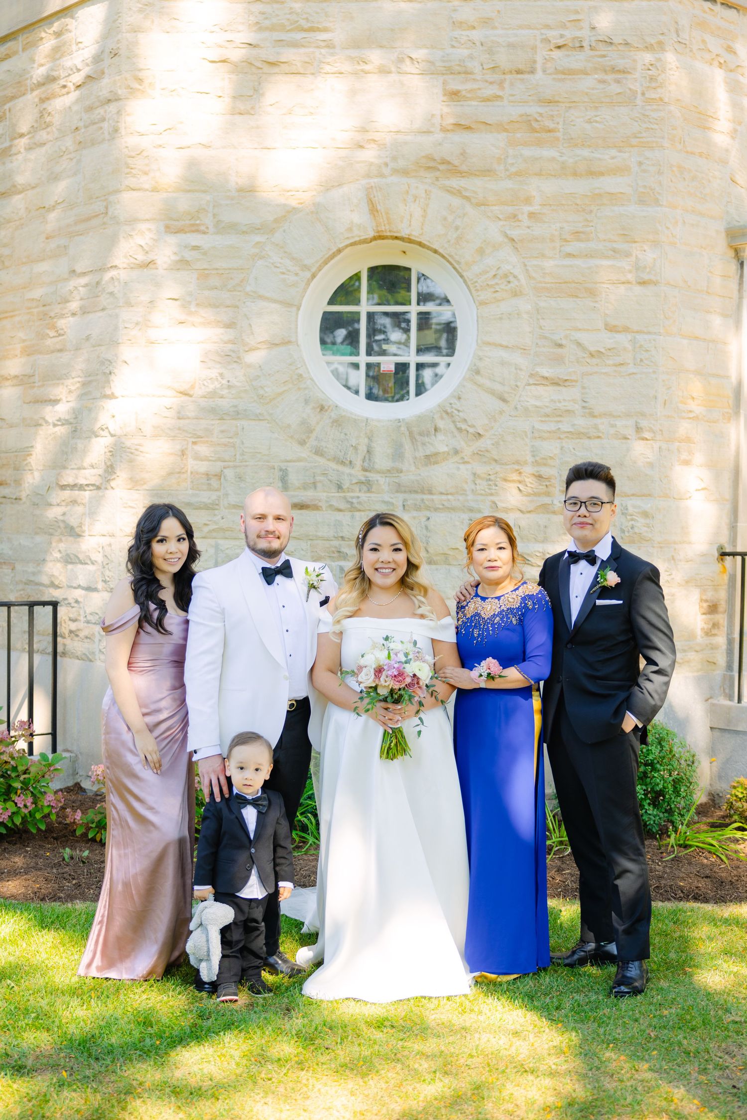 Extended wedding party including small child poses in front of stone church wall with round window.