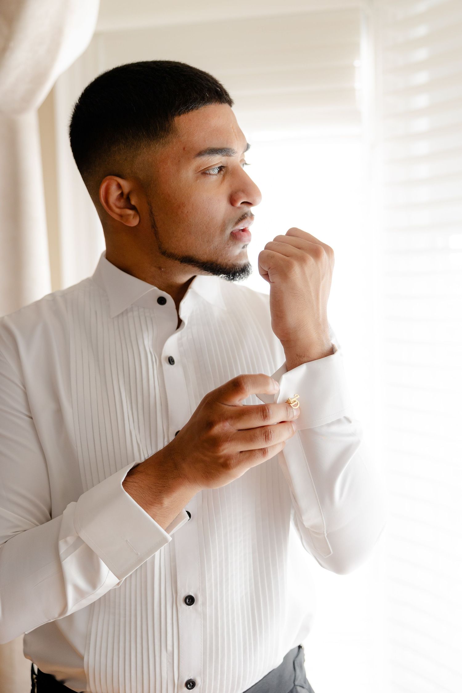 A person in a white pleated shirt adjusts their cuff while standing near a window.