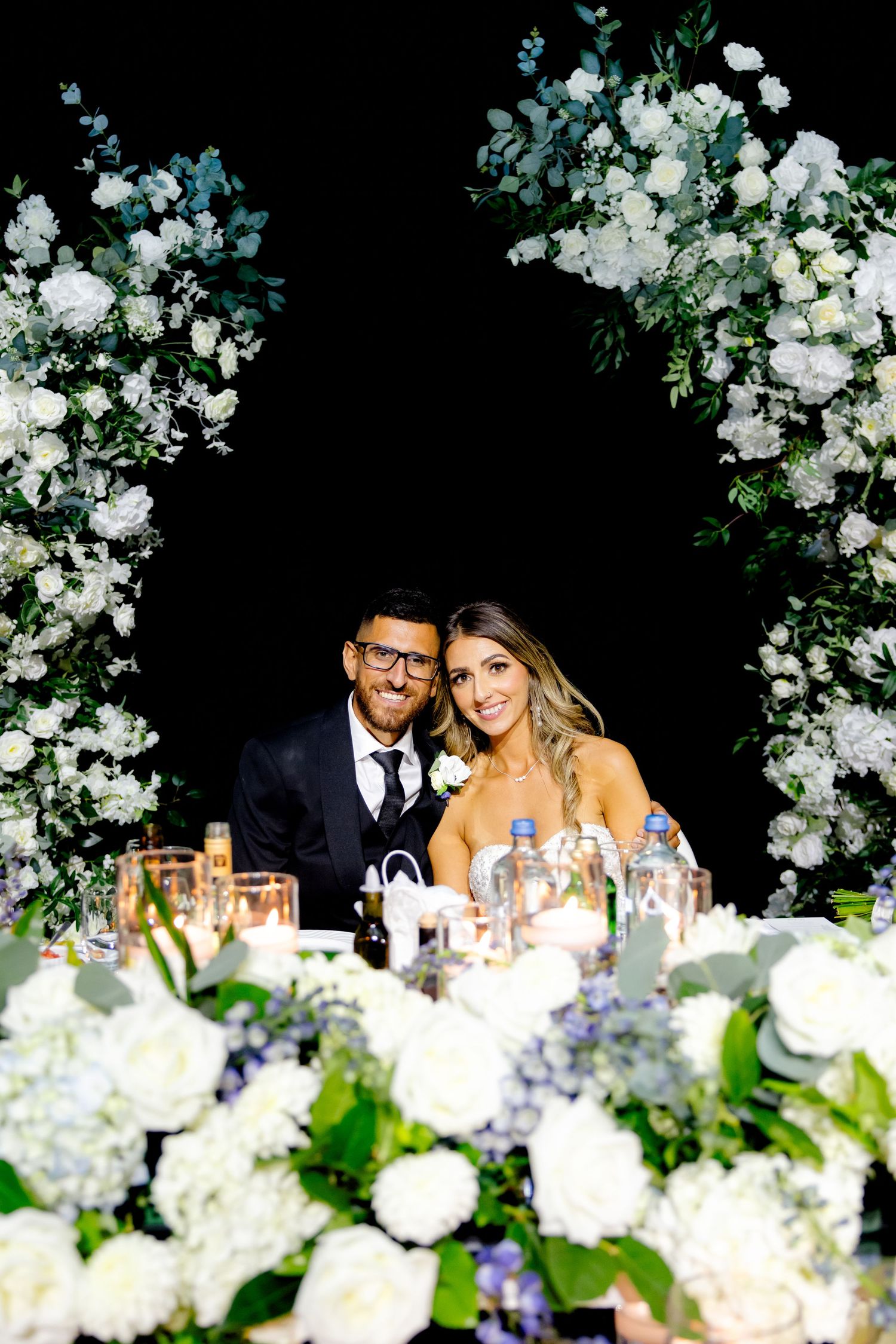 Elegant wedding couple sits behind a stunning floral arch with white and blue blooms against a black background.
