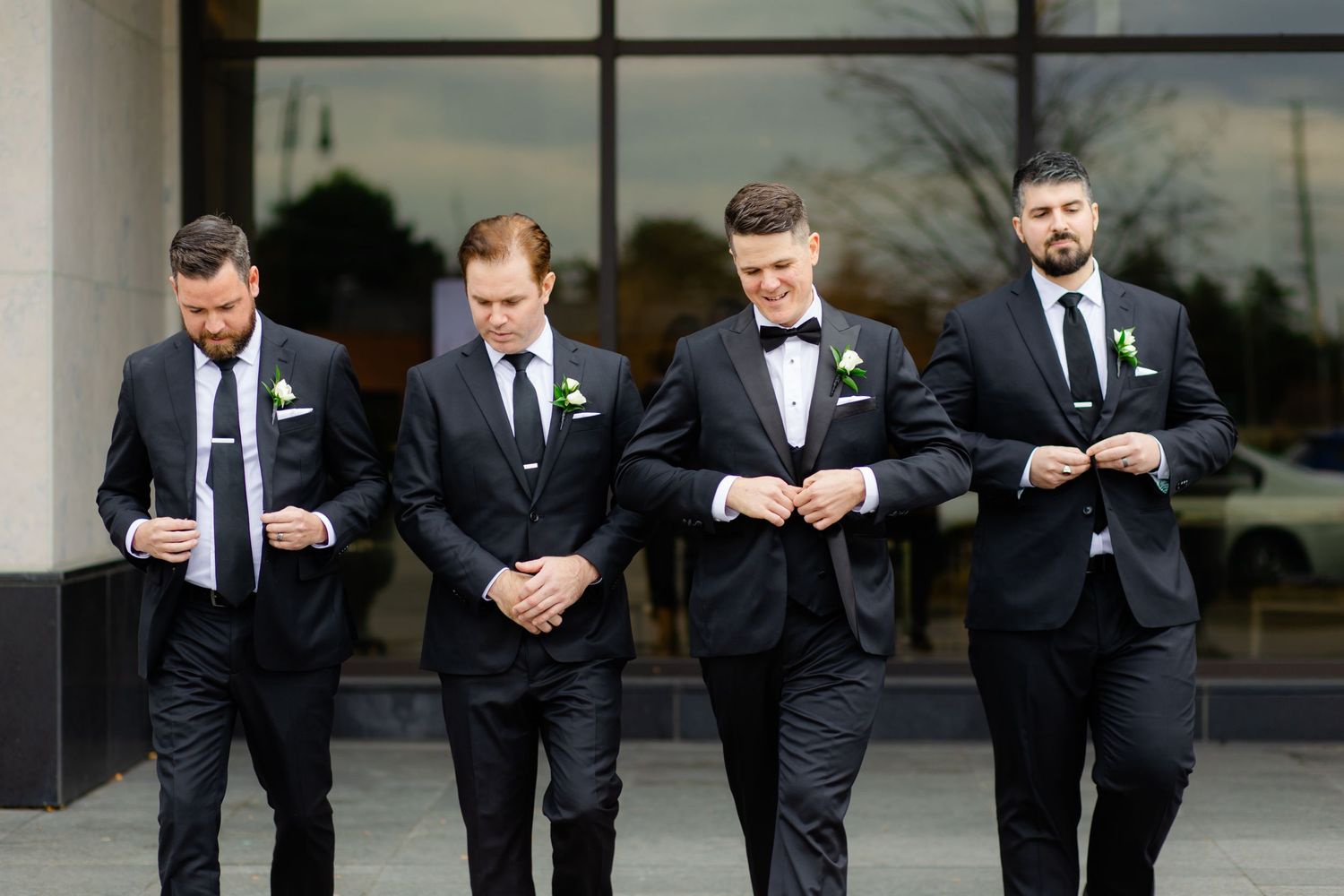 Wedding party groomsmen in formal black tuxedos and bowties stand together outside modern building.