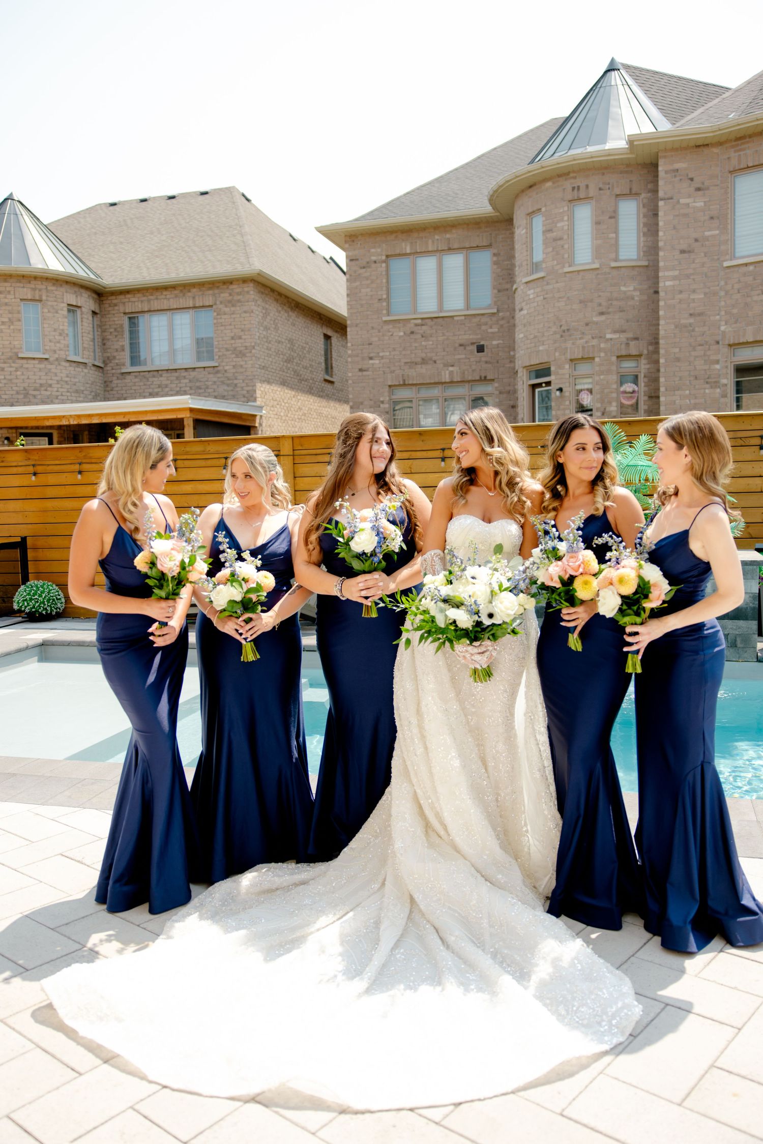 Wedding party with bridesmaids in navy gowns holding bouquets beside a pool.