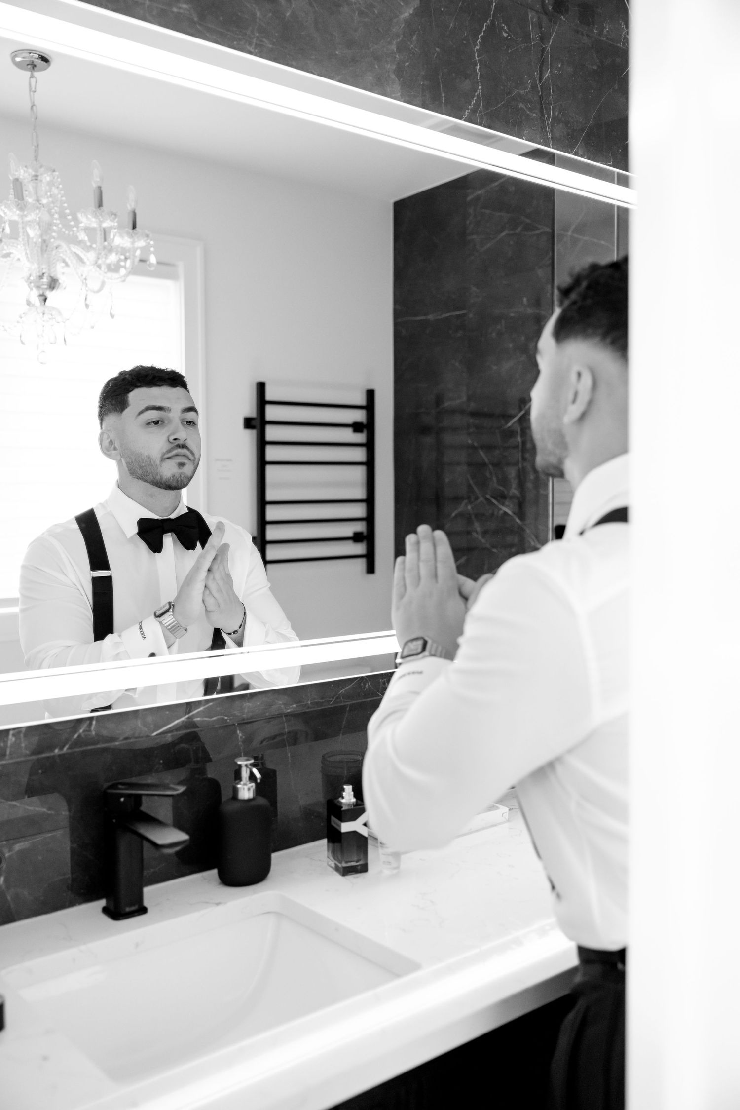 Groom adjusts his bow tie while looking in a modern bathroom mirror with sleek black fixtures and a crystal chandelier.