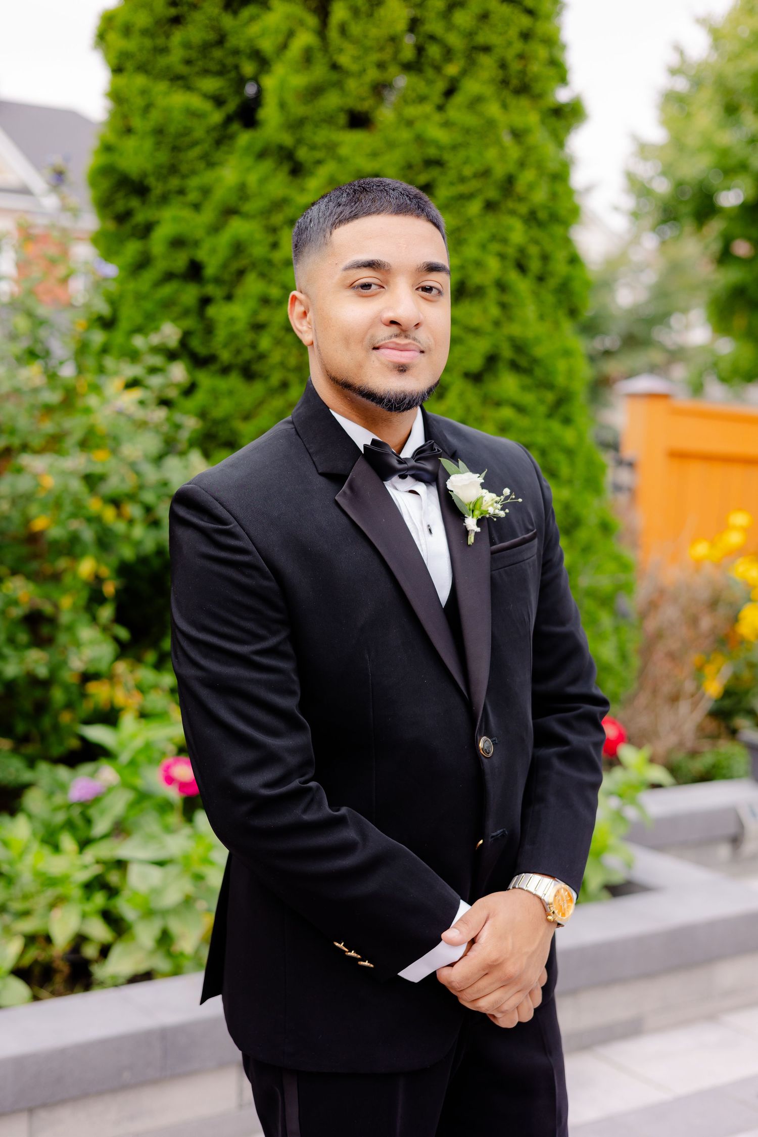 Groom in classic black tuxedo poses in garden setting with greenery backdrop.