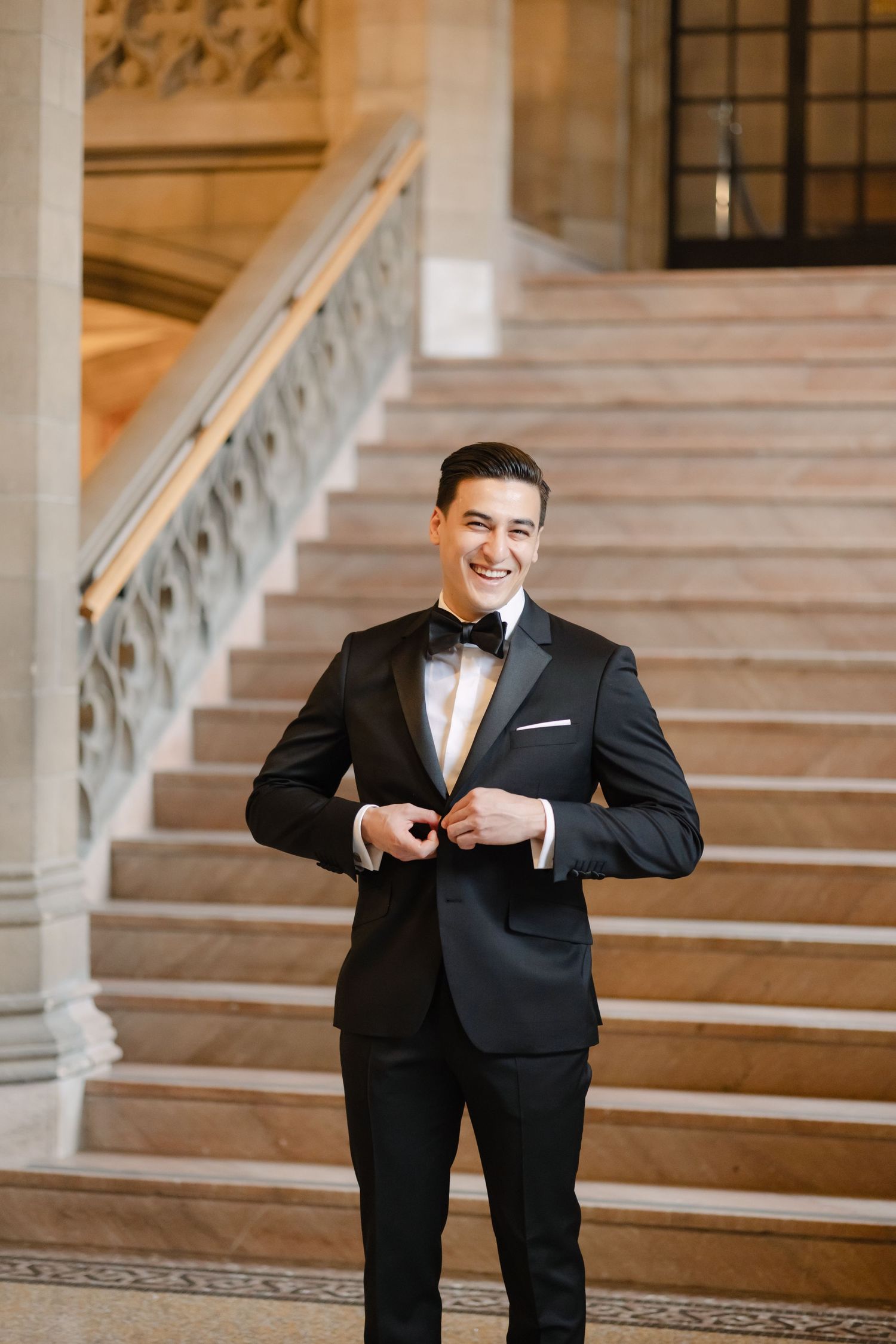 A formal portrait of someone in a black tuxedo and bowtie adjusting their jacket on elegant marble stairs.